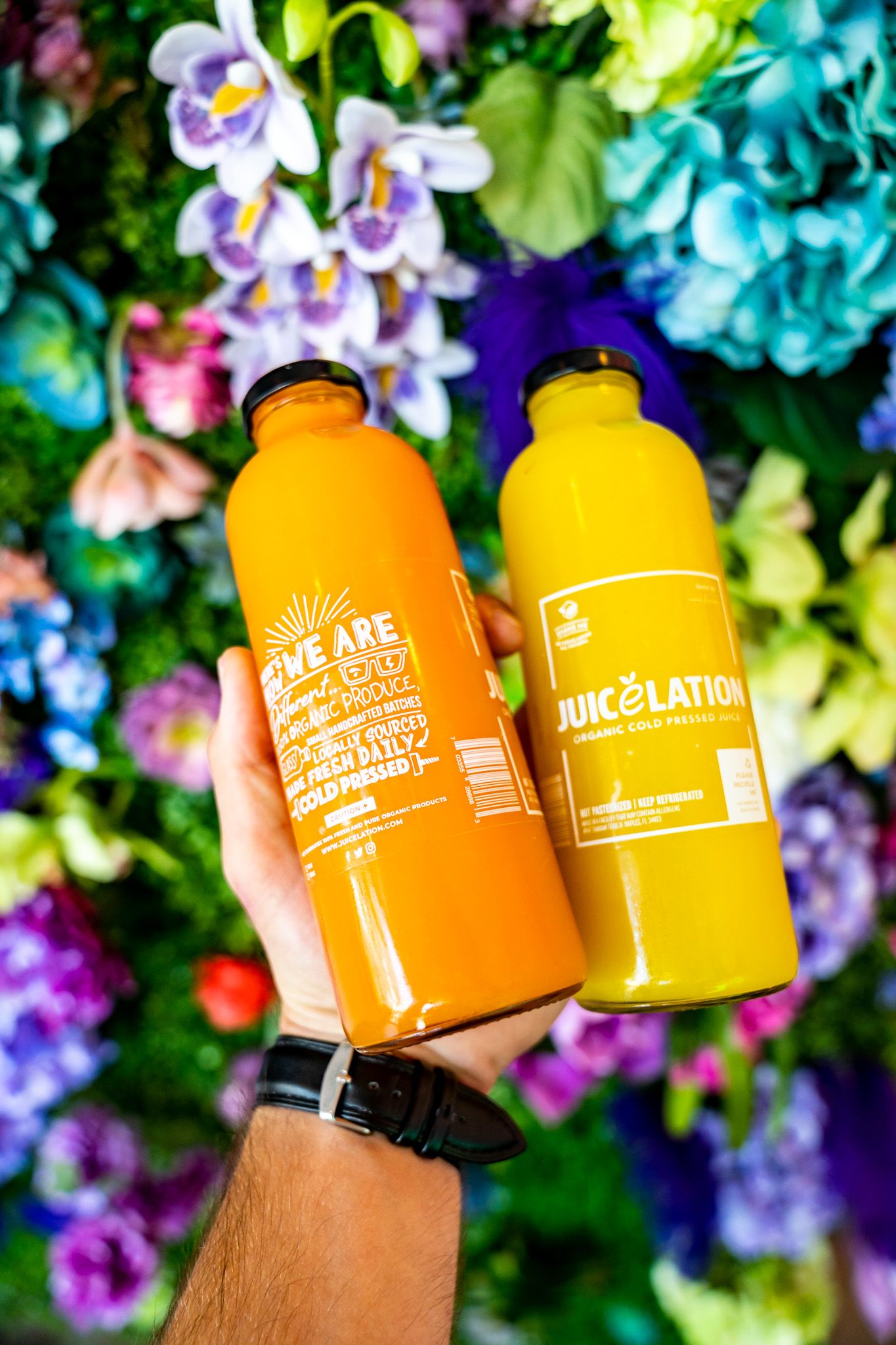 Two bright orange and yellow drink containers held against a colorful floral garden background.