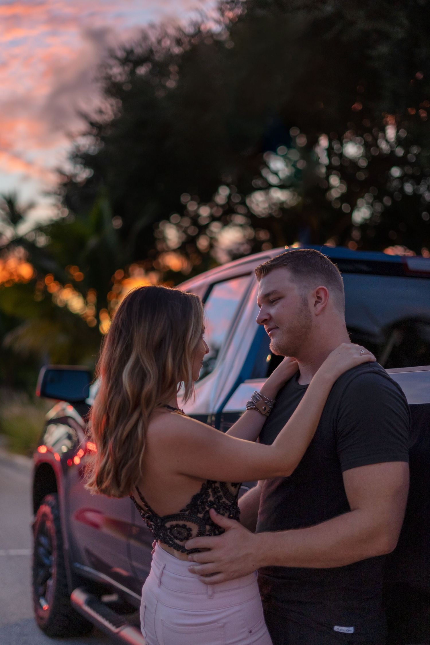 A romantic couple embraces during sunset near parked cars in an intimate moment.