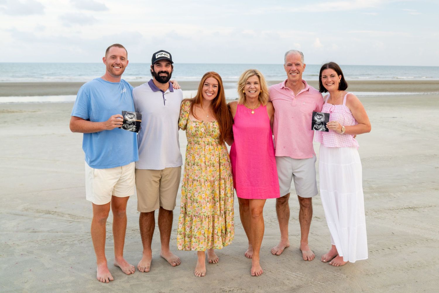 A group in summer attire poses together on a sandy beach during sunset.