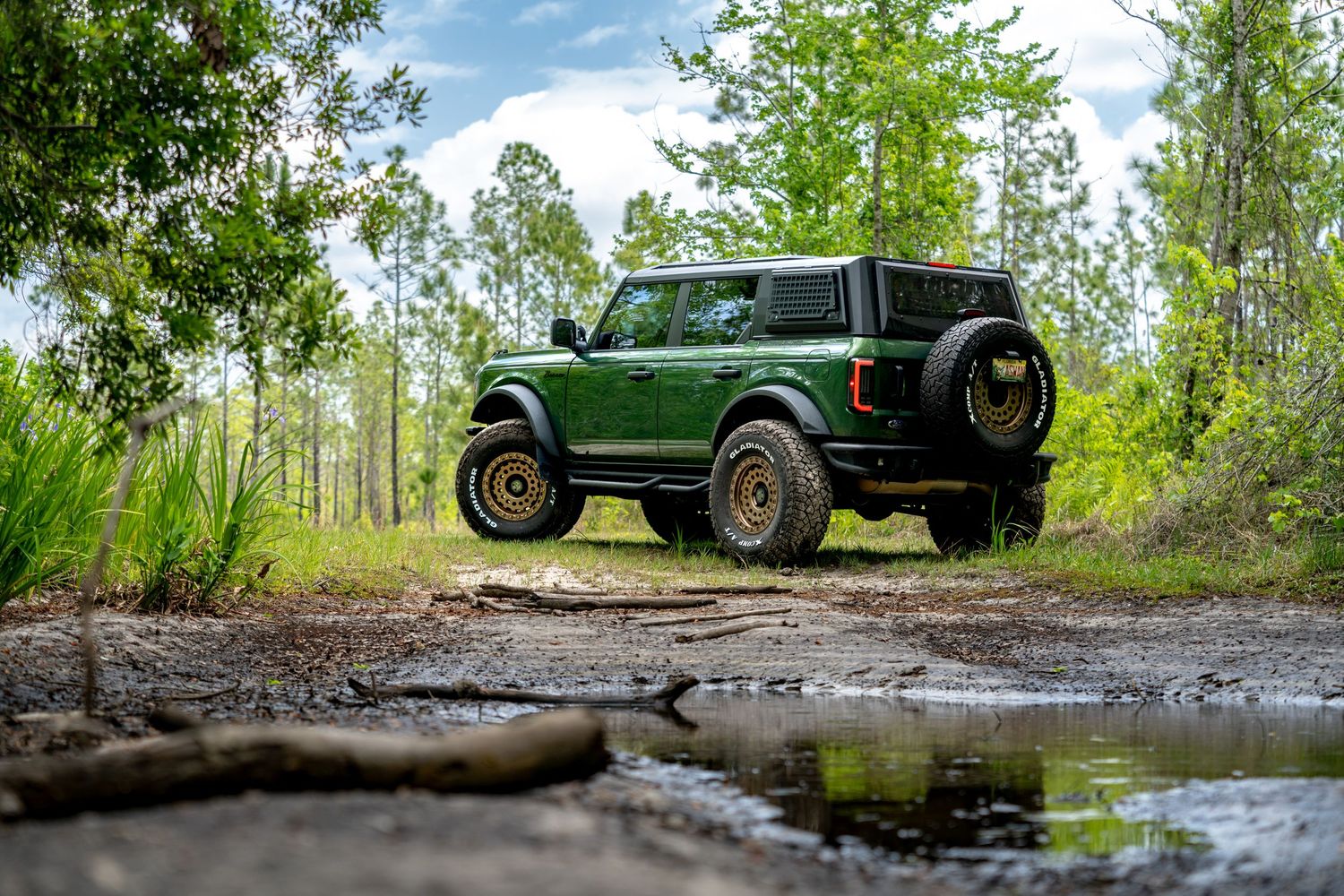 Green Ford Bronco off-road vehicle parked near a muddy puddle surrounded by tall trees in a forest setting.