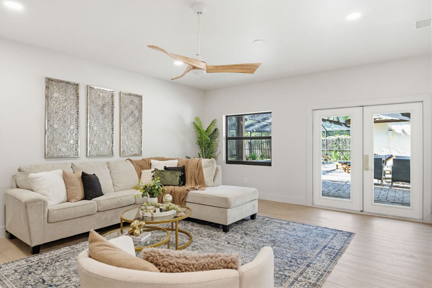 A bright modern living room with beige sectional sofa, ceiling fan, and blue area rug features large windows for natural light.