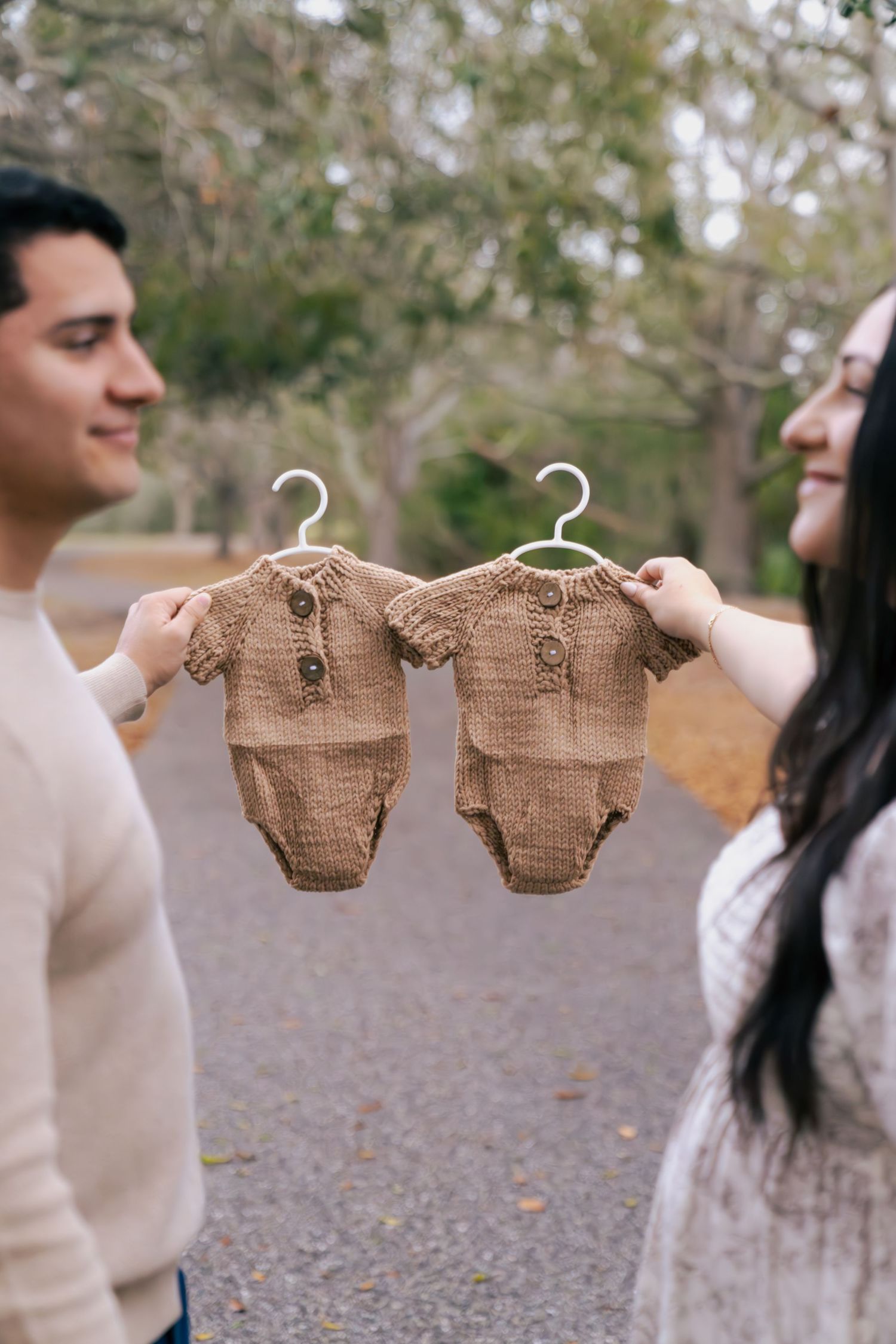 Couple holding up matching knitted brown baby onesies against a blurred outdoor background.
