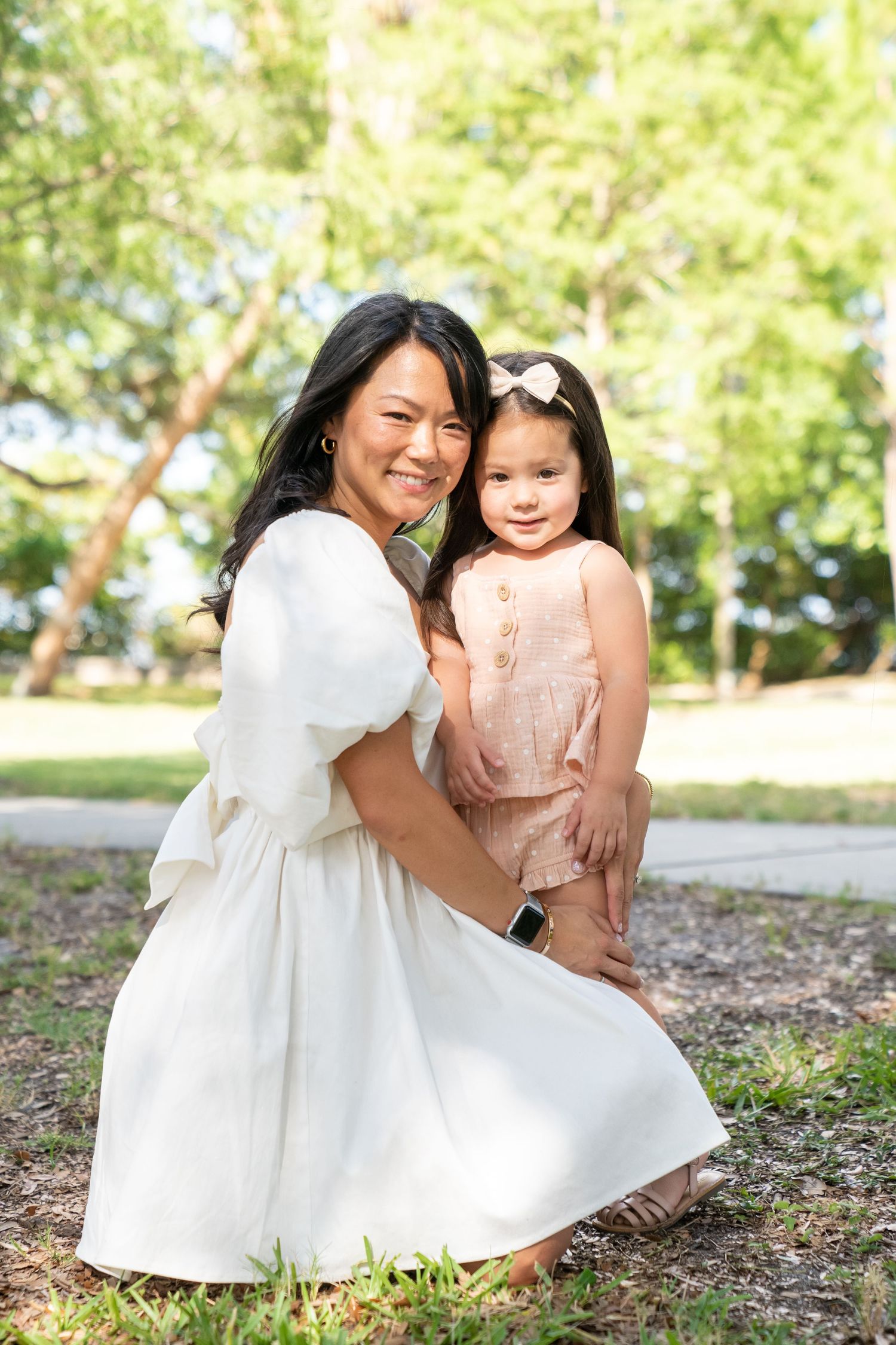 Two people in white and pink dresses sit together on the ground outdoors in a natural garden setting.