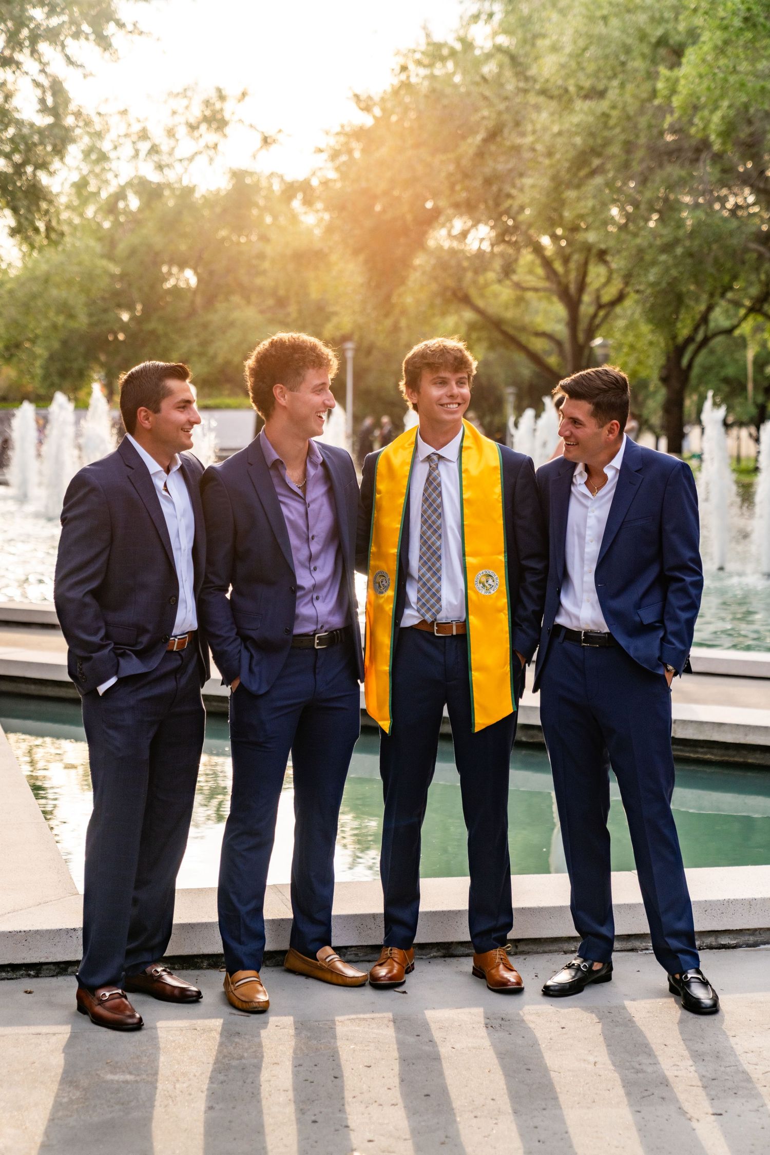 A group of graduates in navy suits and yellow stoles lean against a white wall during golden hour at sunset.