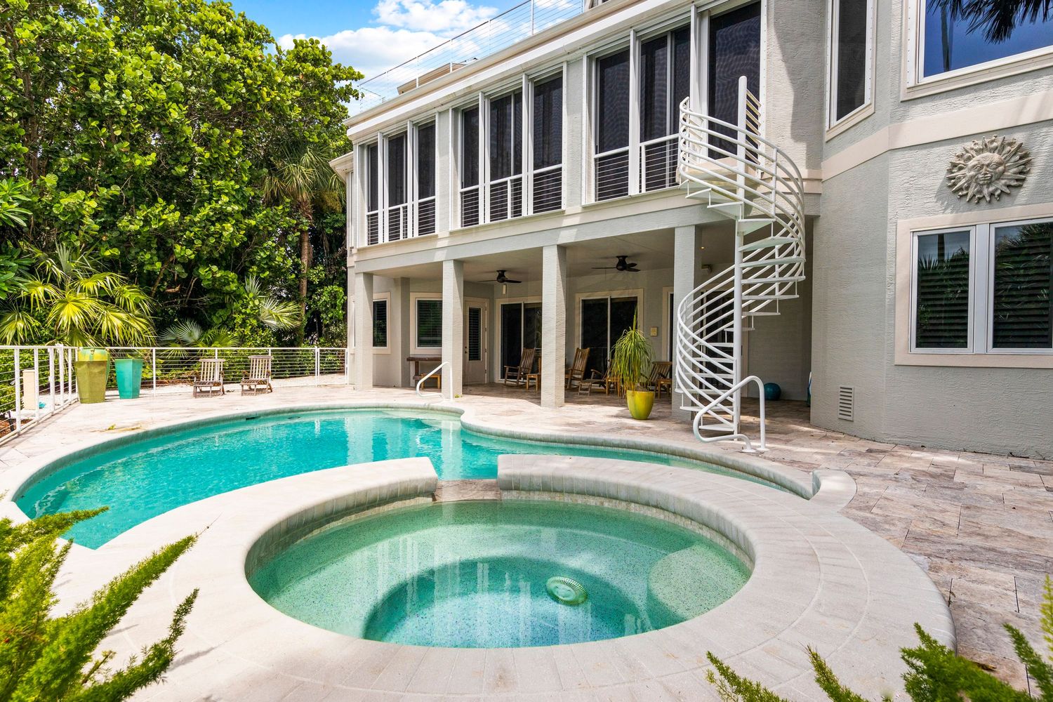 Modern luxury home exterior with curved swimming pool, hot tub and spiral staircase on stone patio surrounded by greenery.