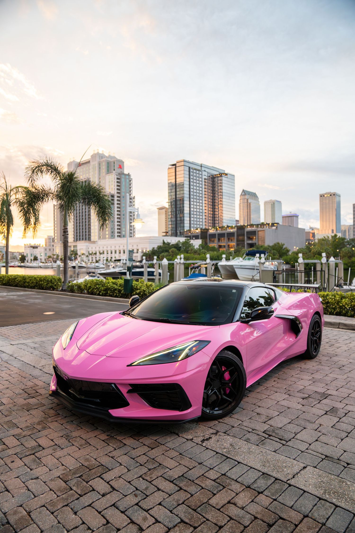 A bright pink McLaren sports car parked on brick pavement with city buildings and palm trees in the background.