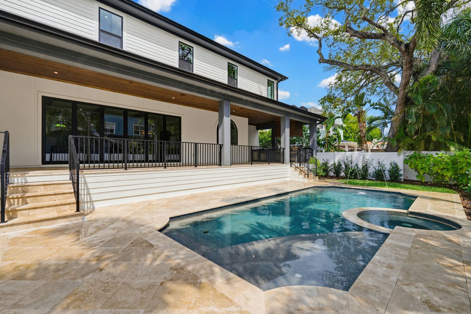 Modern backyard pool area with travertine pavers, glass fencing, and lush landscaping creates a luxurious outdoor oasis.