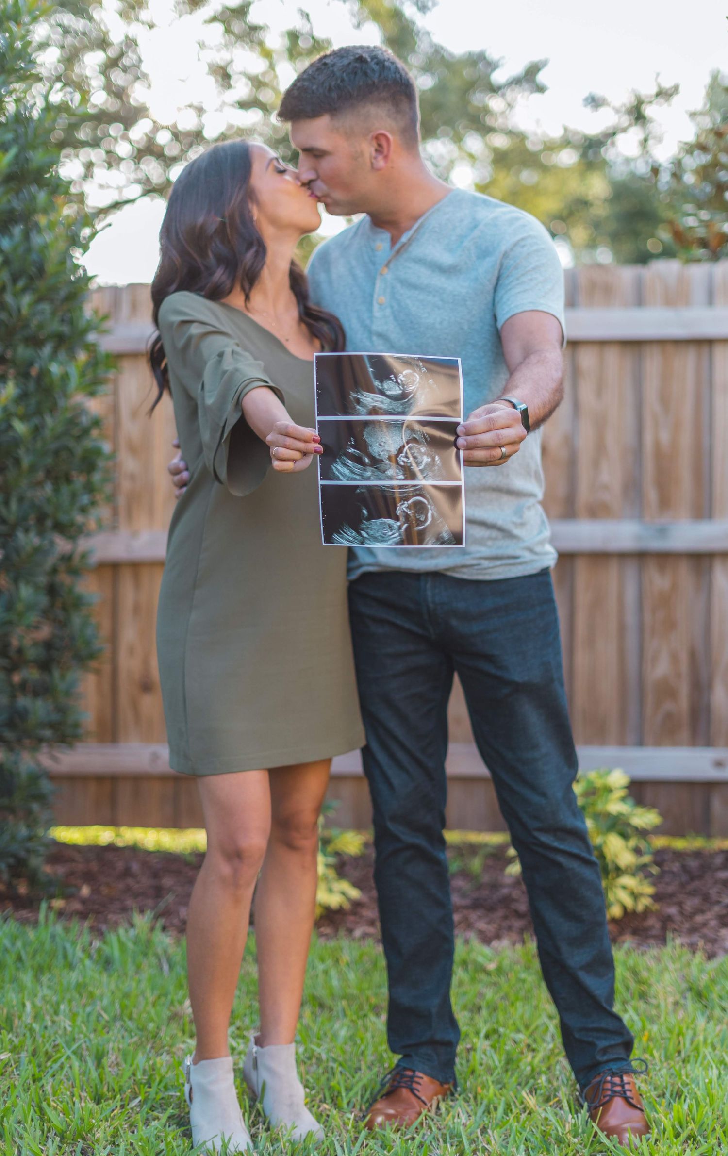 A couple stands in a backyard garden holding ultrasound photos while sharing a tender moment together.