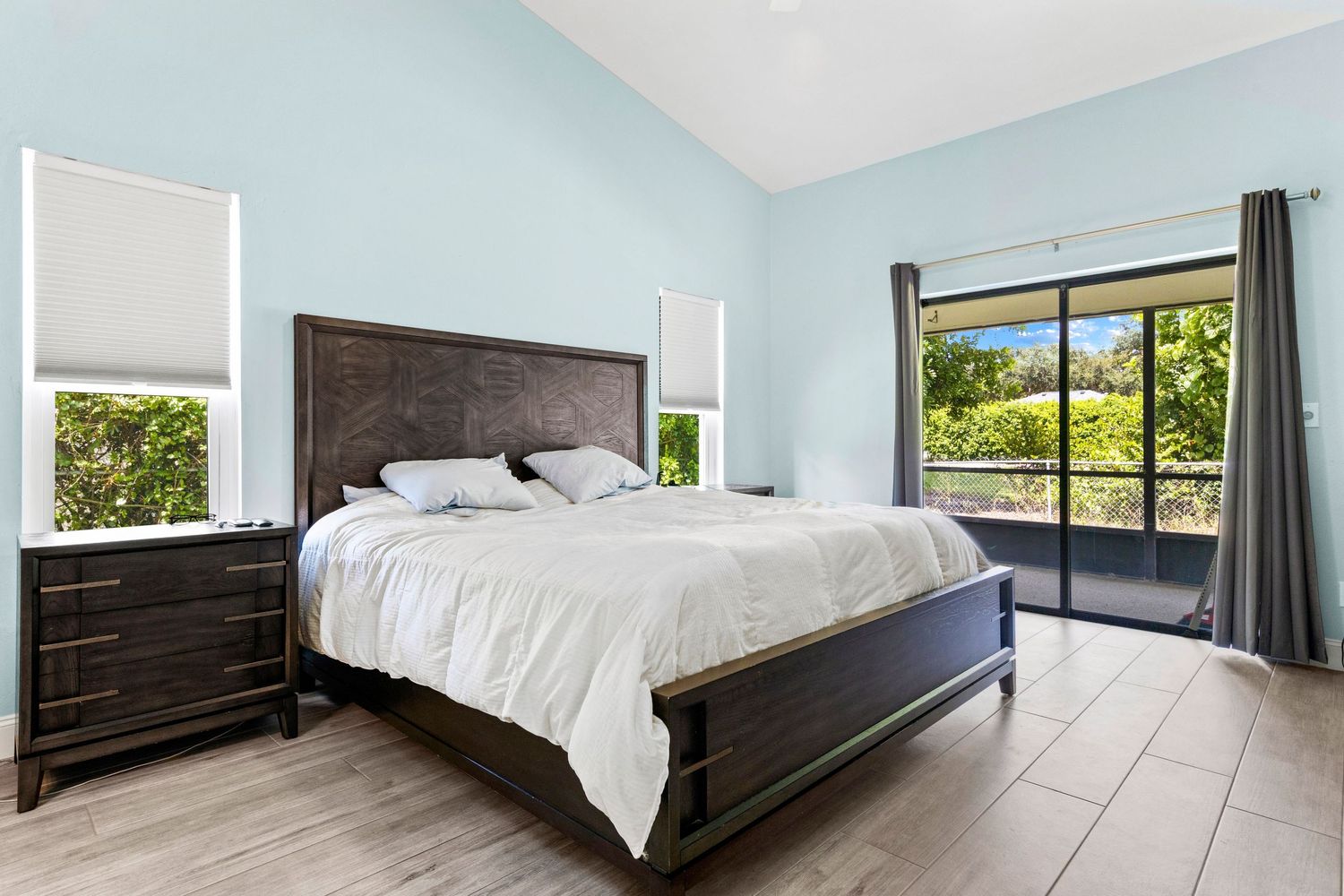 Modern bedroom interior with white bedding on dark wood frame and large windows letting in natural light.