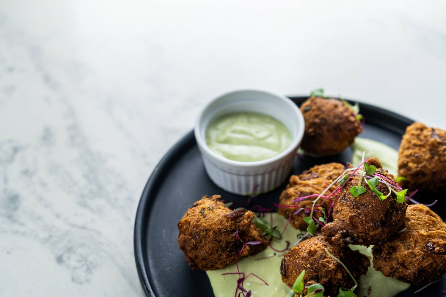 Crispy golden fried croquettes served on a dark plate with creamy green dipping sauce and fresh herbs.