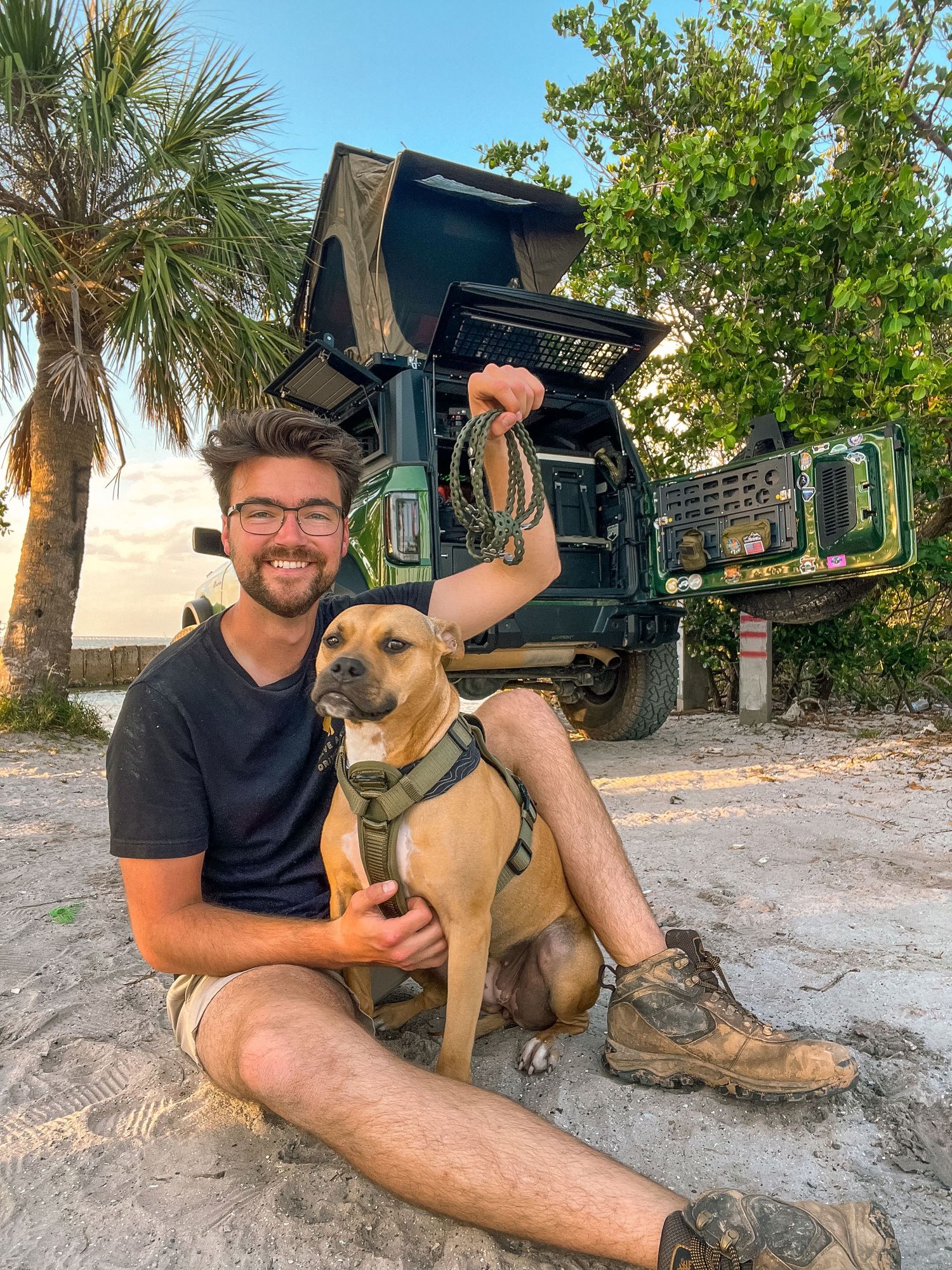 A brown dog sits with its owner on sandy ground surrounded by palm trees and greenery.