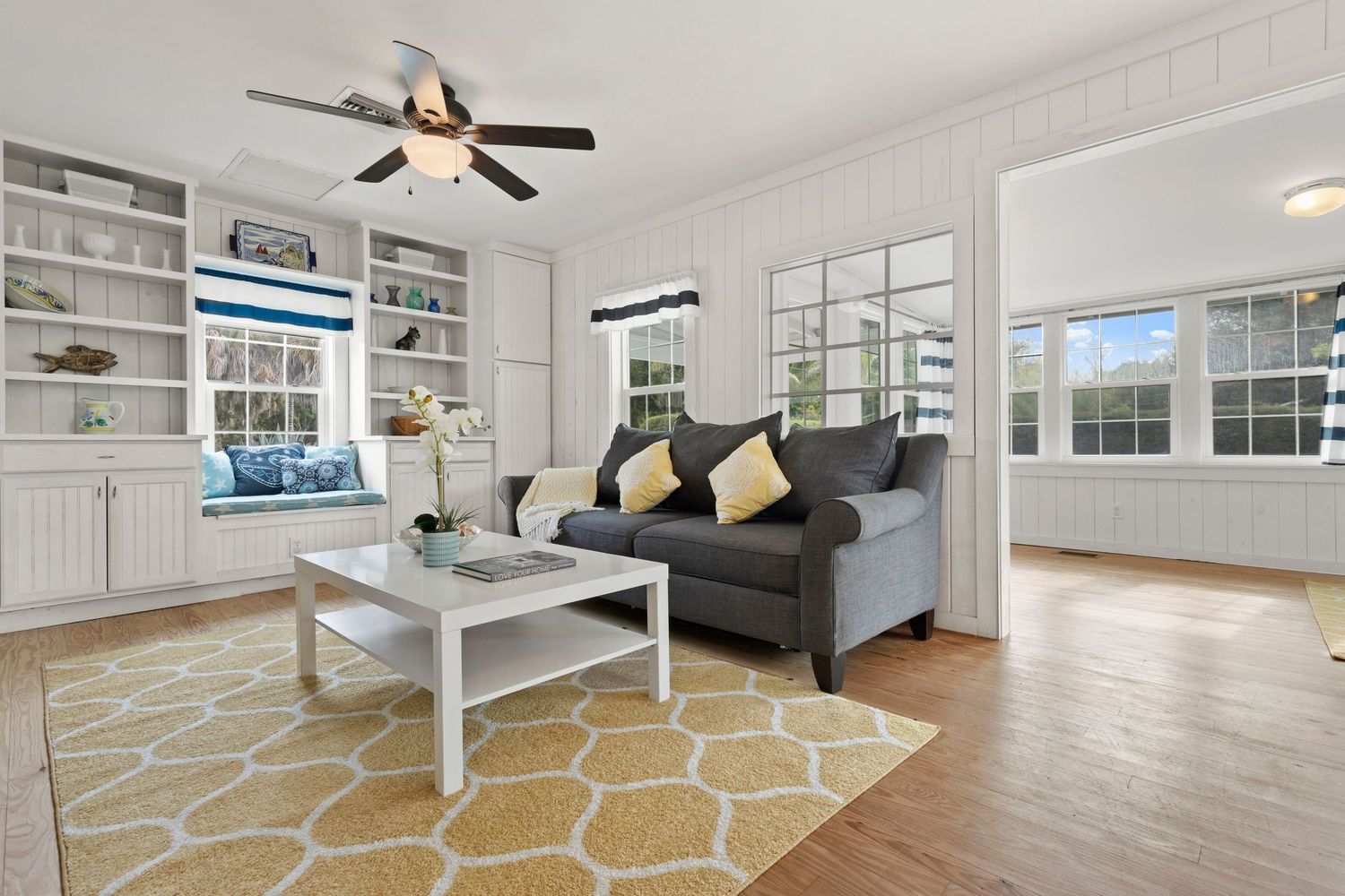 Modern living room with ceiling fan, built-in shelving, and hardwood floors showcasing bright natural lighting.