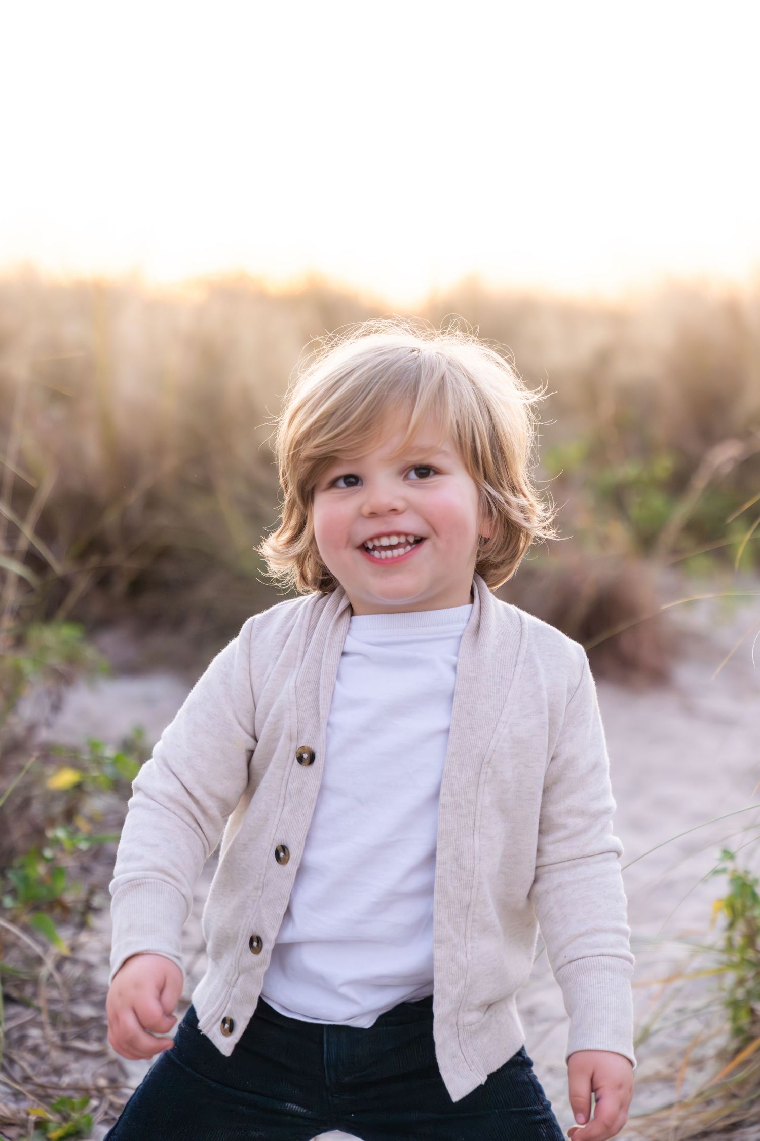 A child in a beige cardigan and white shirt sits outdoors during golden hour with a joyful expression.