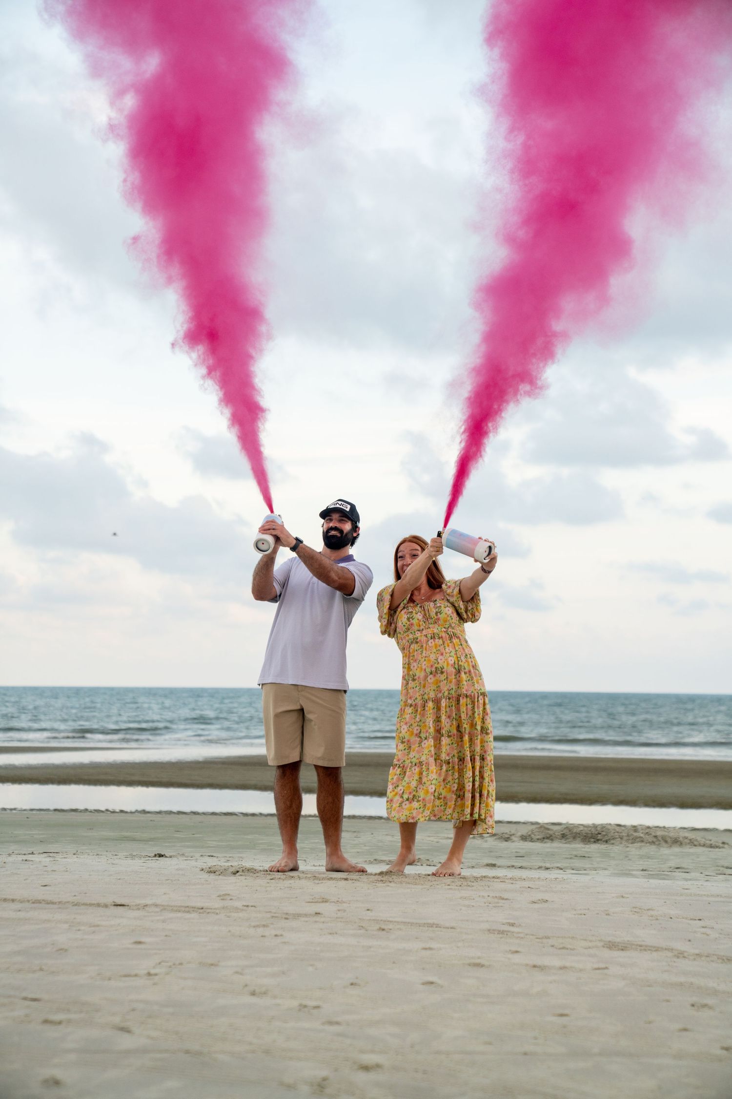 A couple on a beach holds pink smoke flares during a photo shoot with the ocean in the background.