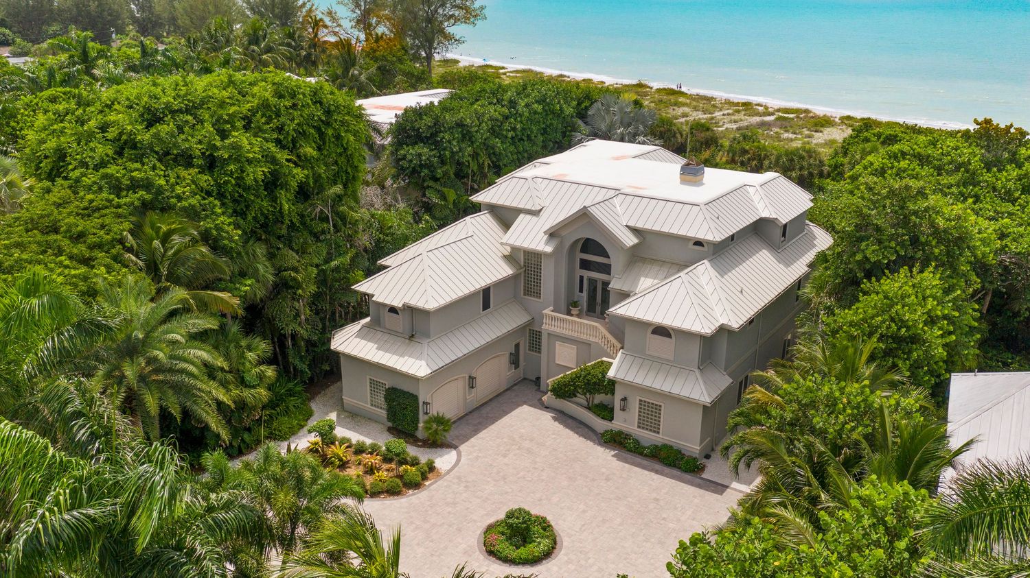Aerial view of luxury beachfront home with white roof and tropical landscaping surrounded by lush palm trees.