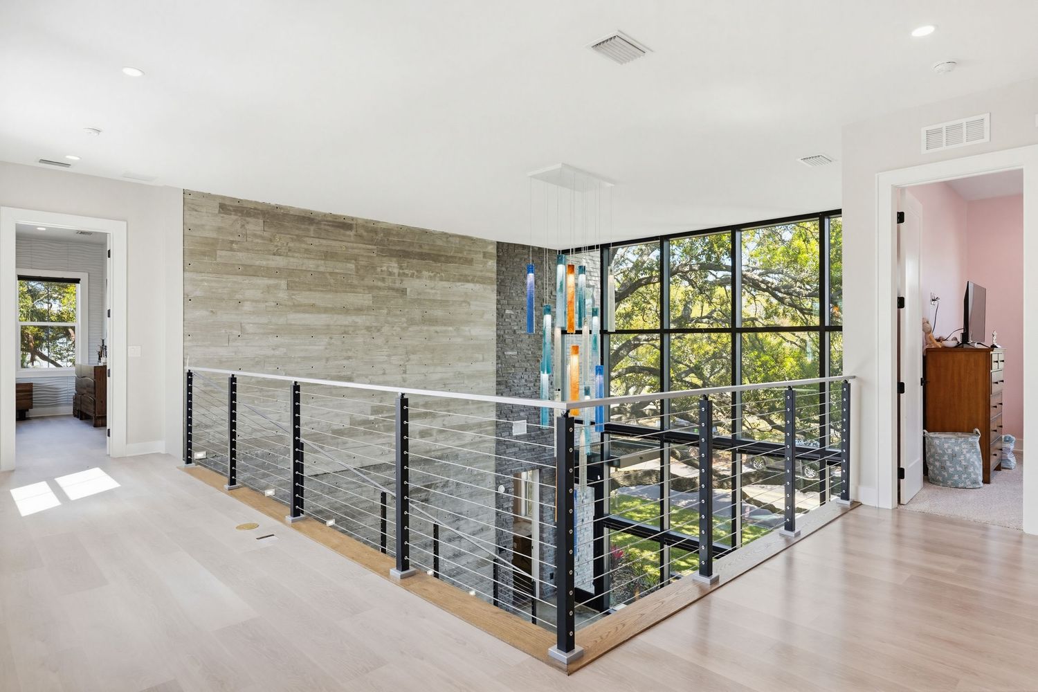 Modern upstairs hallway with light wood floors, black metal railing, and concrete accent wall overlooking windows.