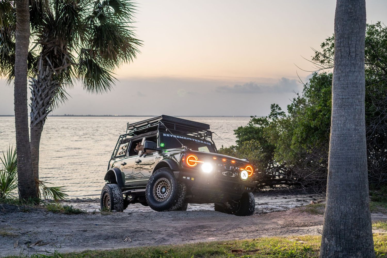 Off-road vehicle silhouetted against sunset by the ocean with palm trees.