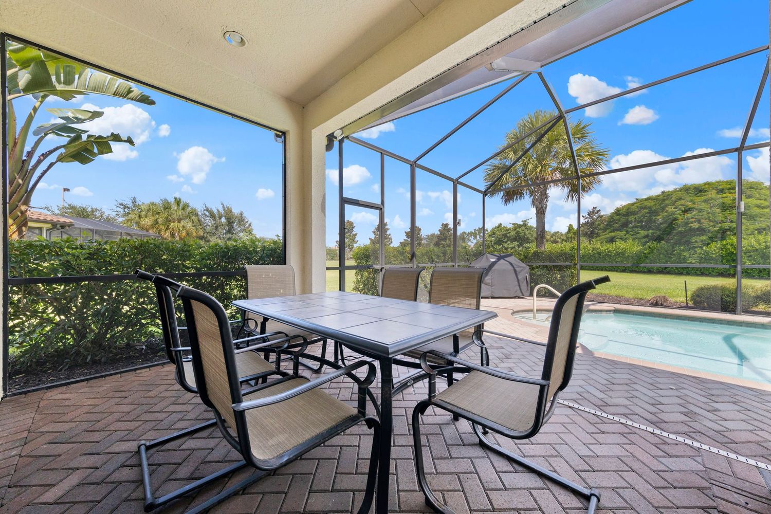 Covered patio with outdoor dining furniture overlooking a swimming pool and tropical landscaping on a sunny day.
