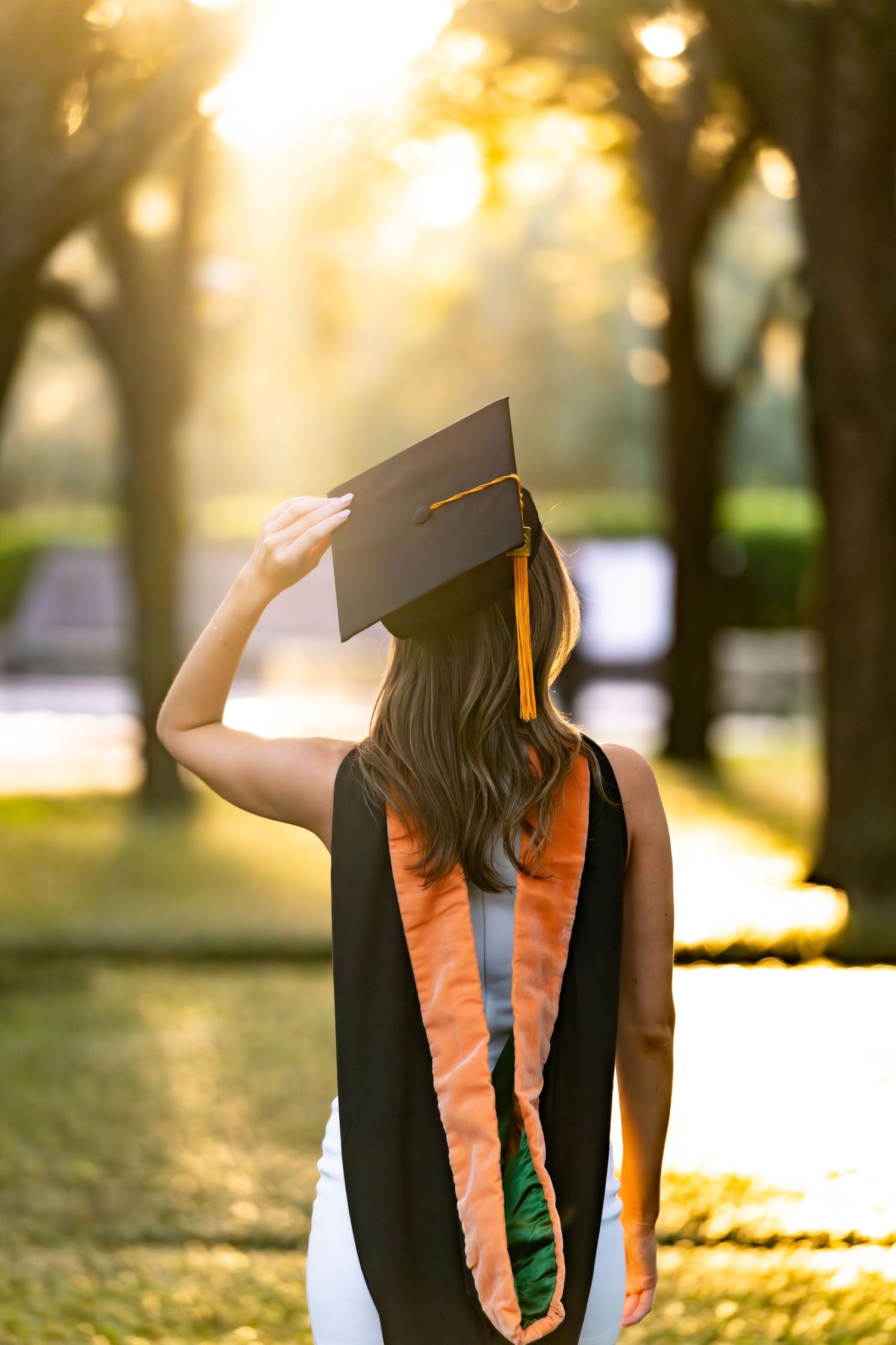 Graduate in cap and gown poses during golden hour sunset in a park with sunlight filtering through trees.