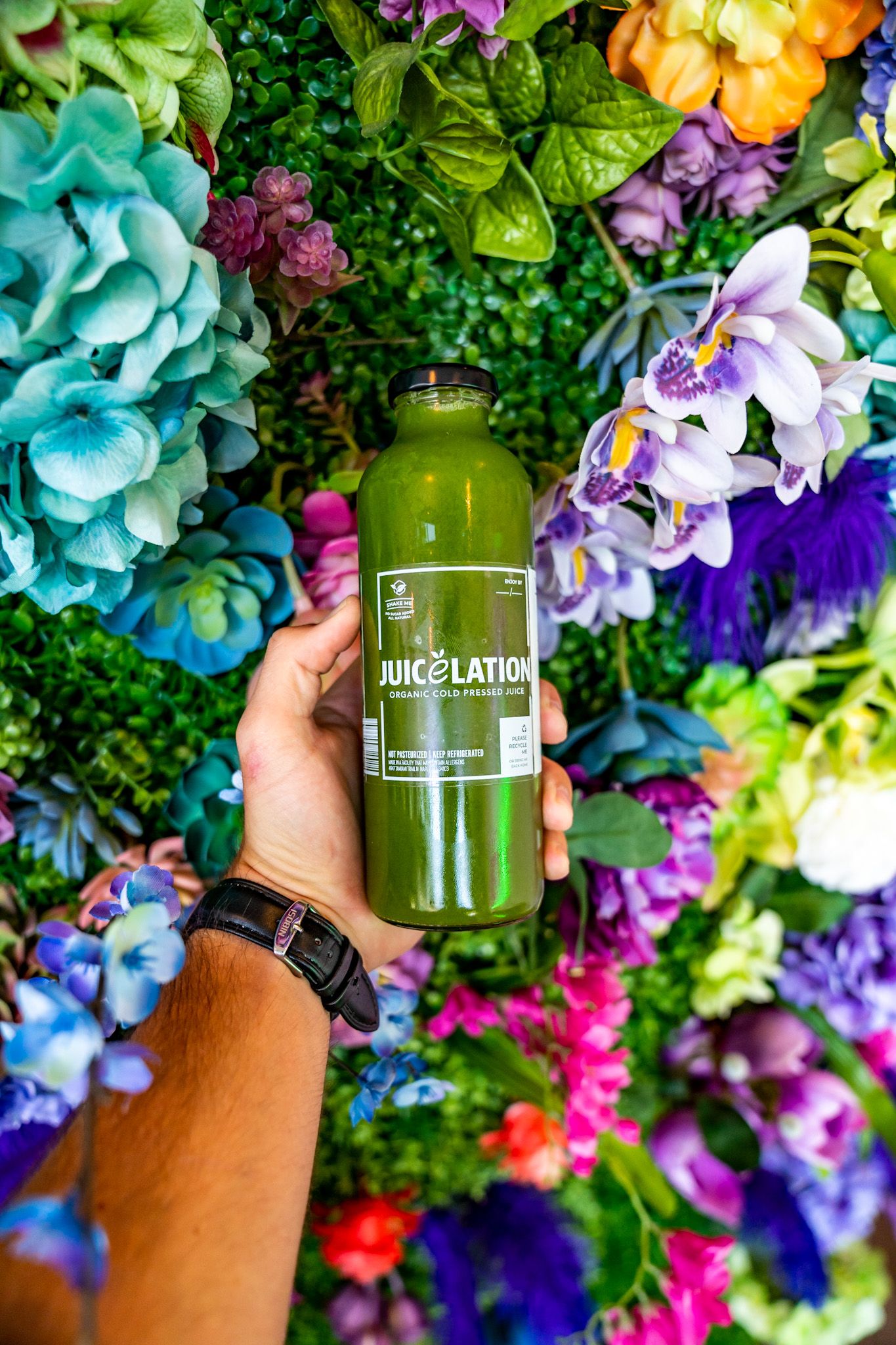 Green juice bottle held against colorful floral backdrop with turquoise and purple artificial flowers.