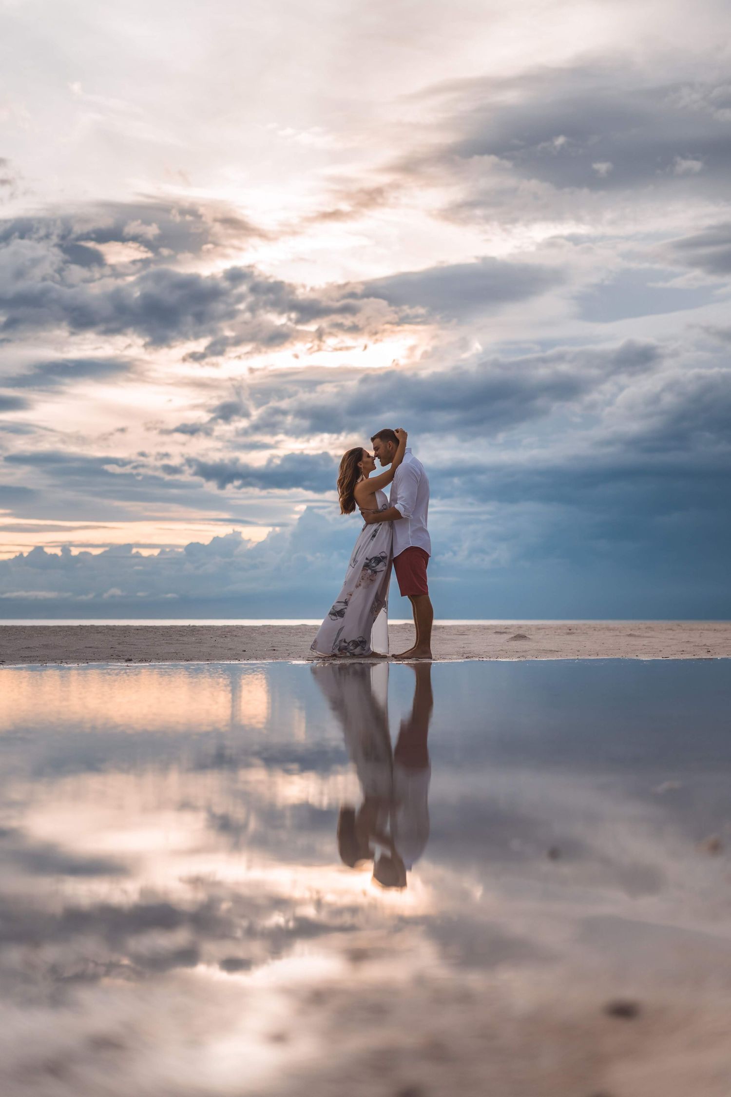 Two figures embrace on a reflective salt flat surface at sunset with dramatic cloudy skies above.