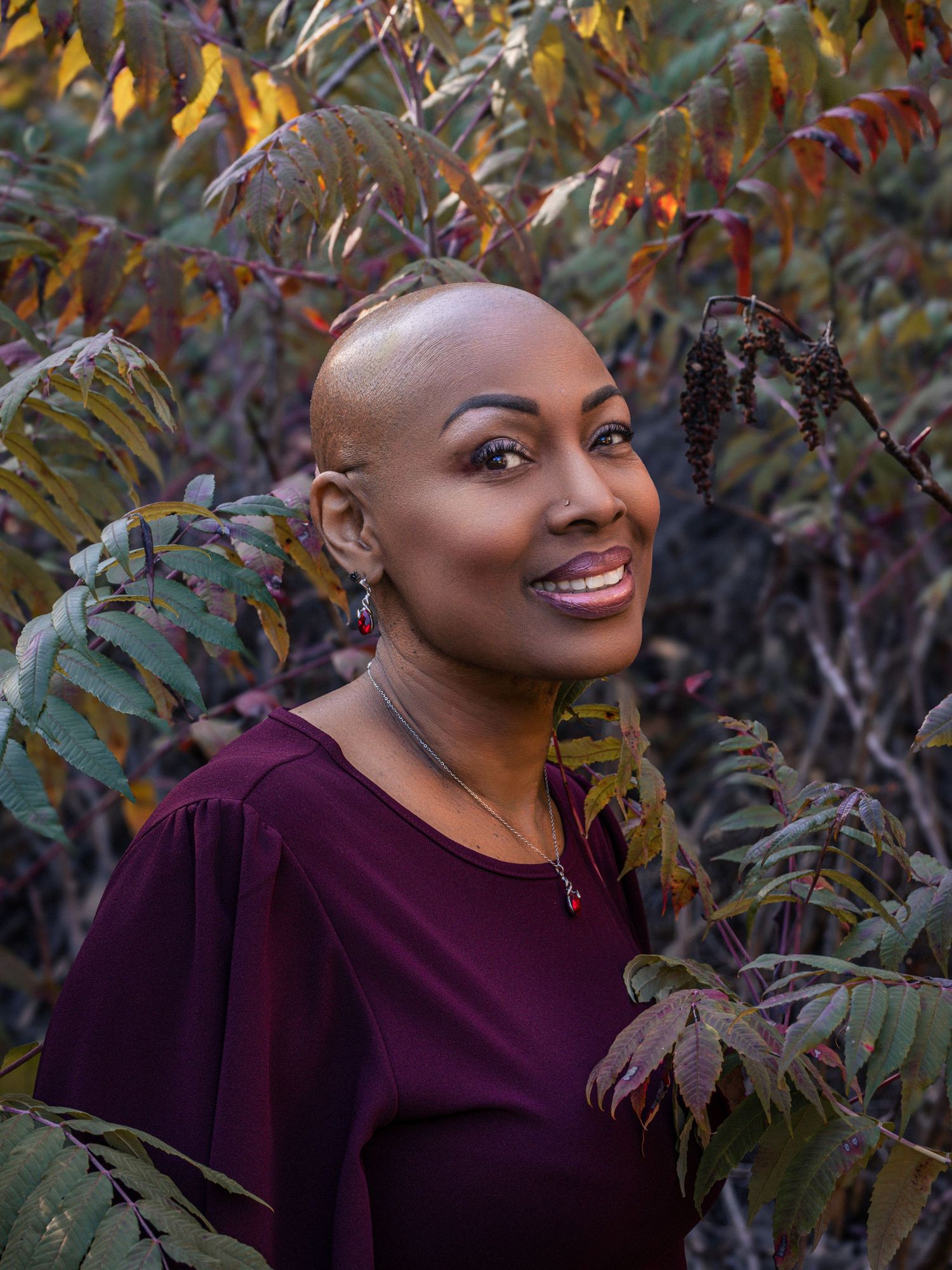 A smiling bald Black woman in a burgundy top poses among colorful autumn foliage in an outdoor portrait.
