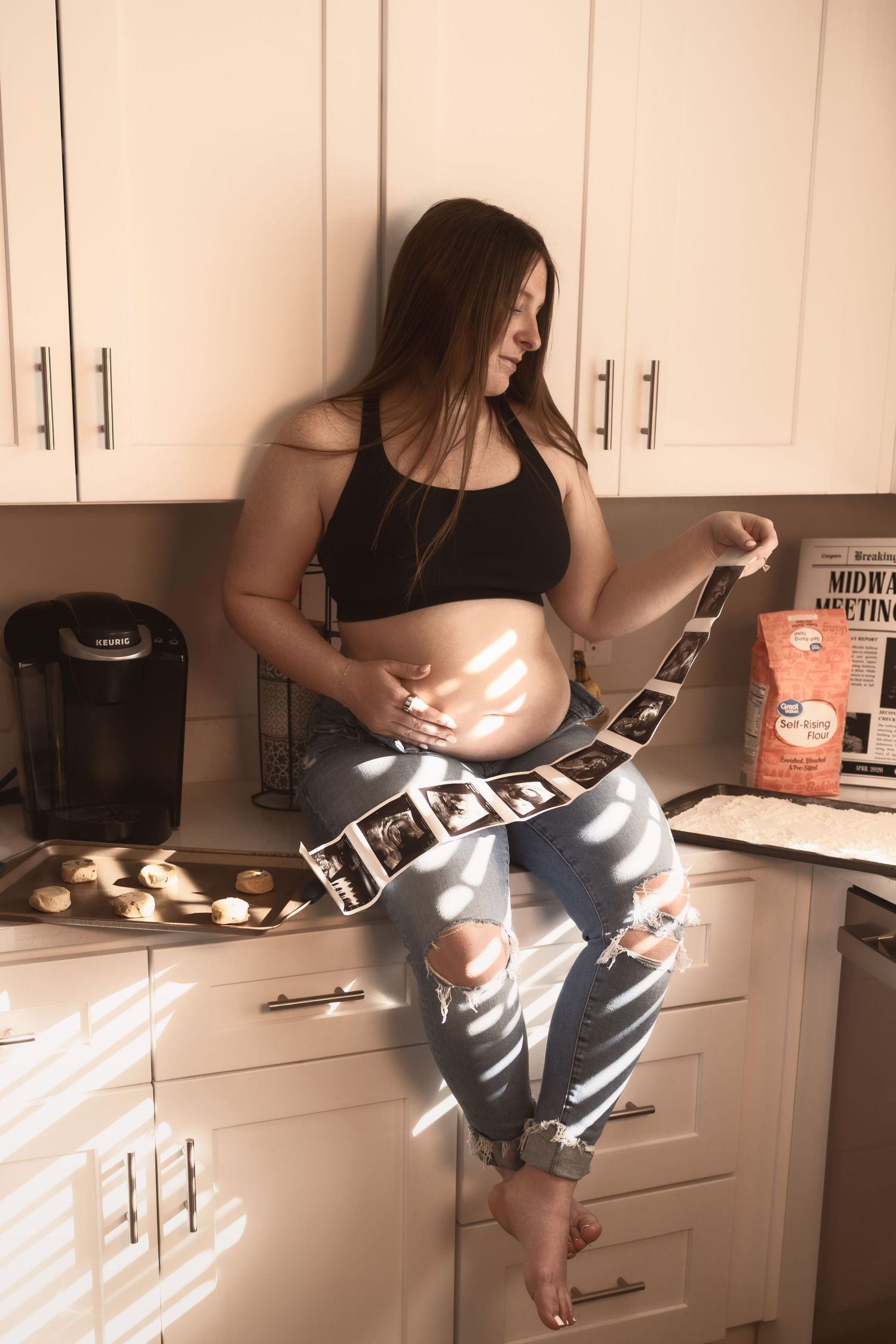 Pregnant woman sitting on kitchen counter in ripped jeans reviewing ultrasound photos in warm sunlight.