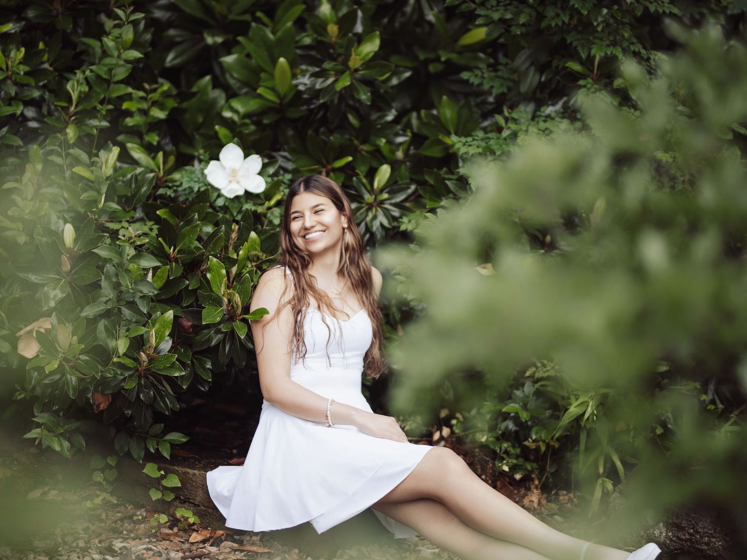 Woman in white dress relaxing among green foliage and flowers.