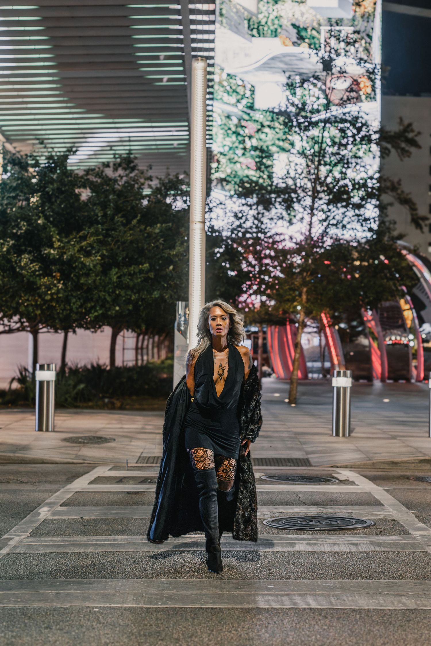 A stylish woman in a black coat and thigh-high boots walks confidently through an urban plaza at night.