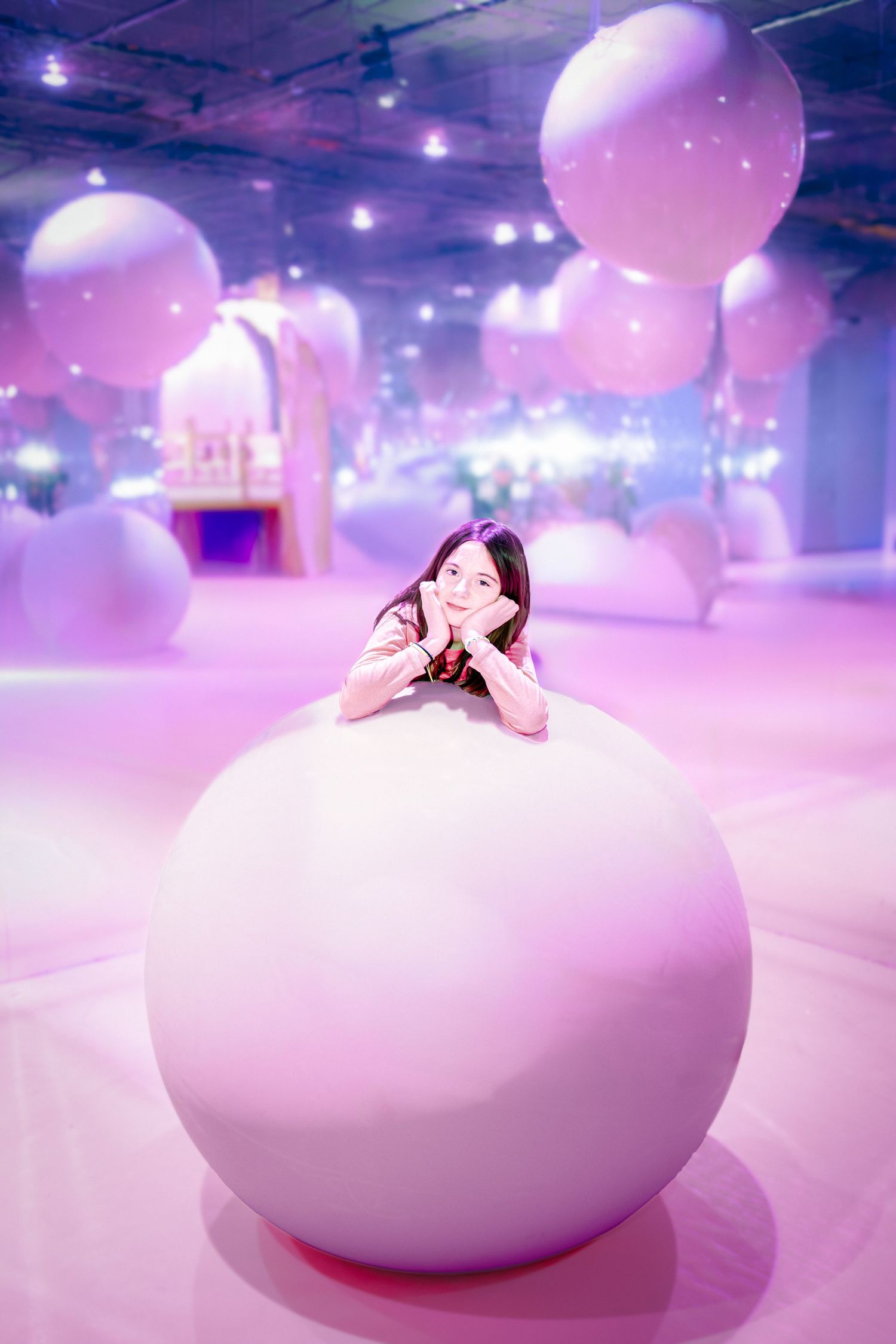 A smiling young woman leans on a large pink ball in a dreamy pink room filled with floating white balloons.