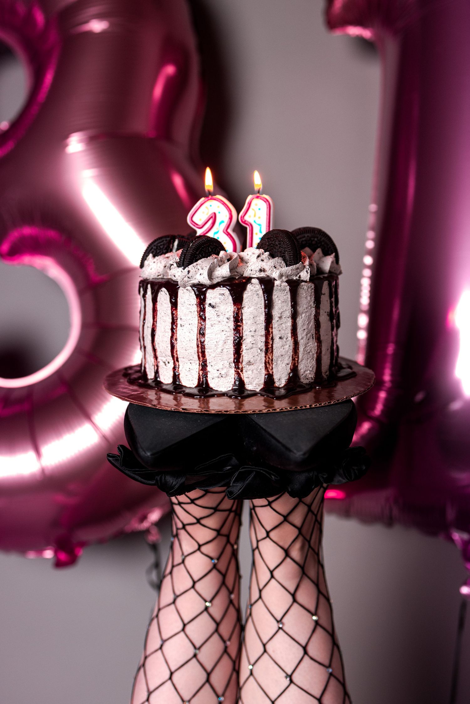 A 31st birthday cake with lit candles held up by fishnet-stocking legs, with pink number balloons in background.
