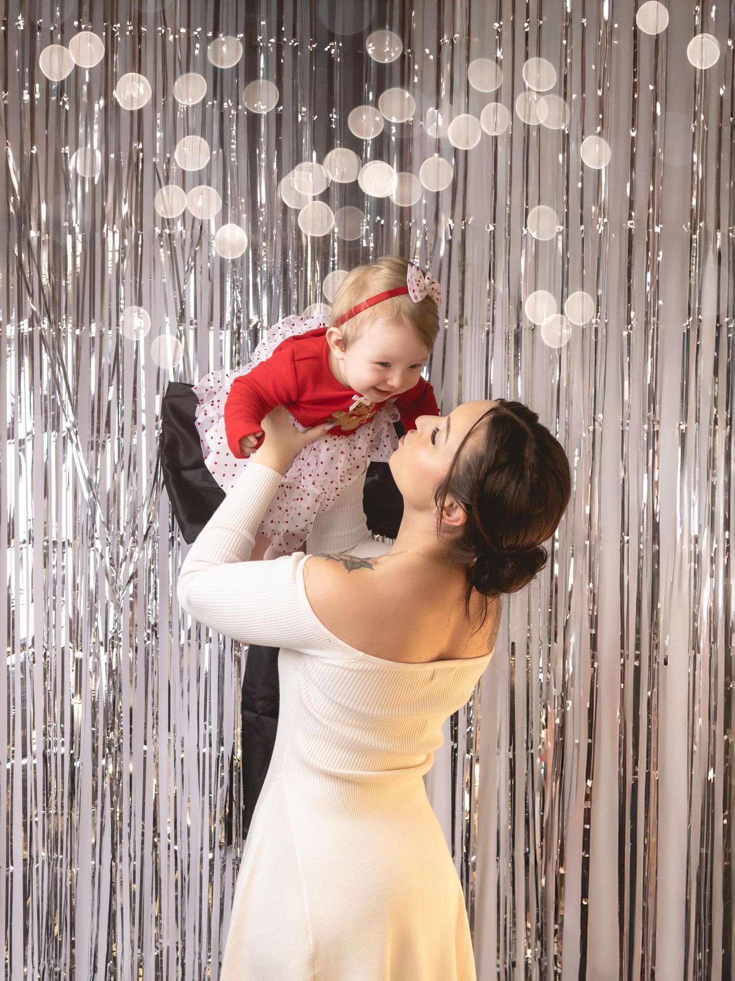 A woman in a white dress lifts a baby in a red outfit in front of a silver tinsel curtain backdrop.