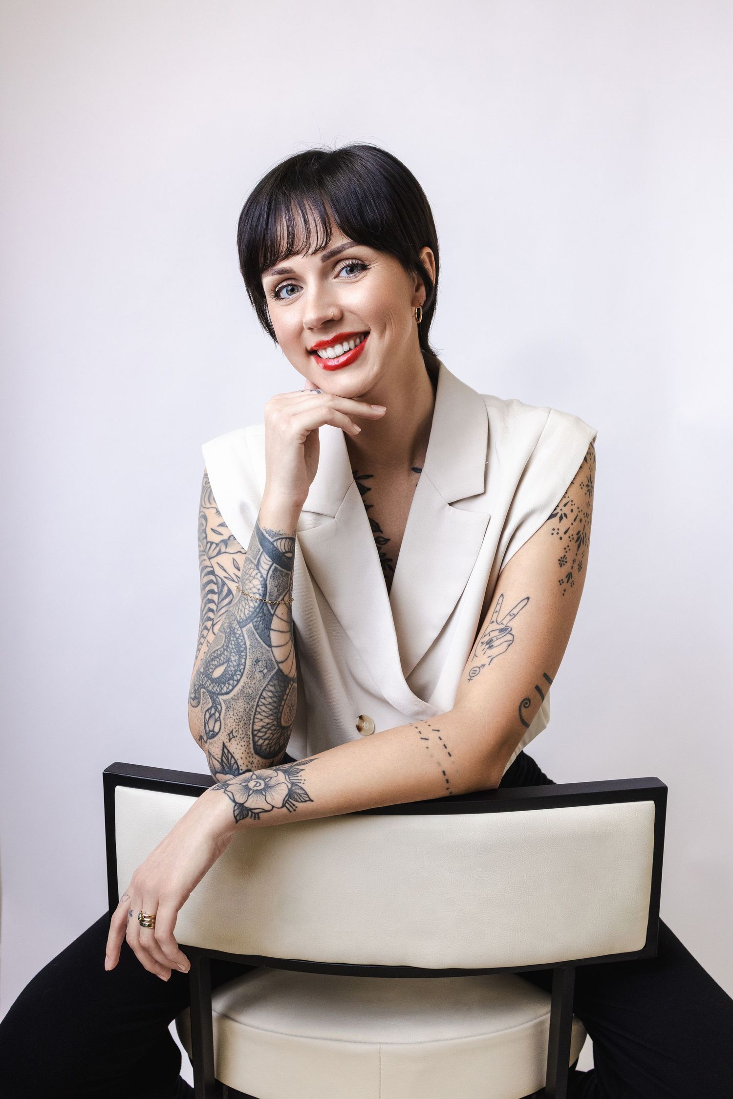 A person in a white blazer with visible arm tattoos poses while seated in a modern cream-colored chair against a light background.