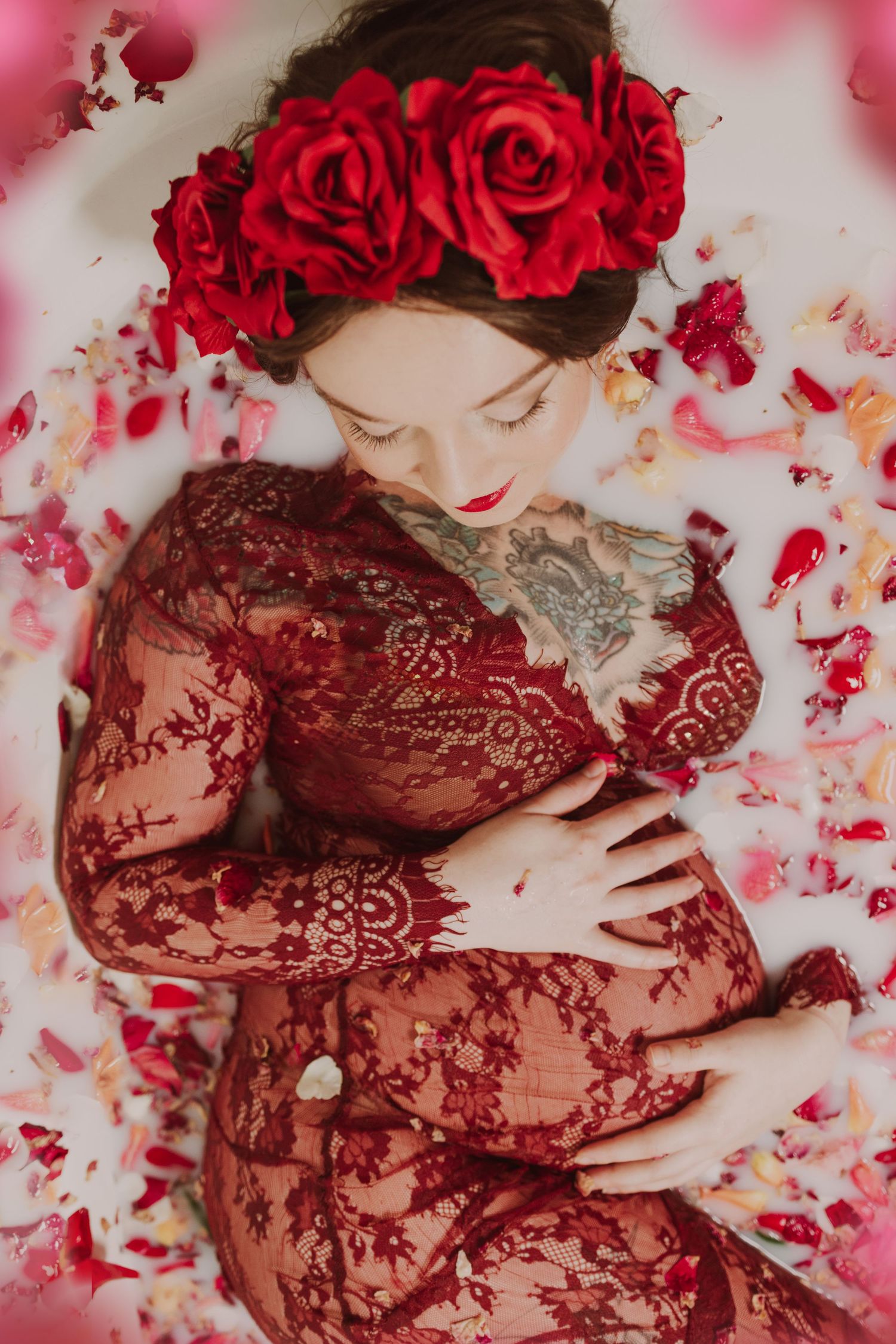 Artistic maternity photo with red roses and scattered petals on white background.