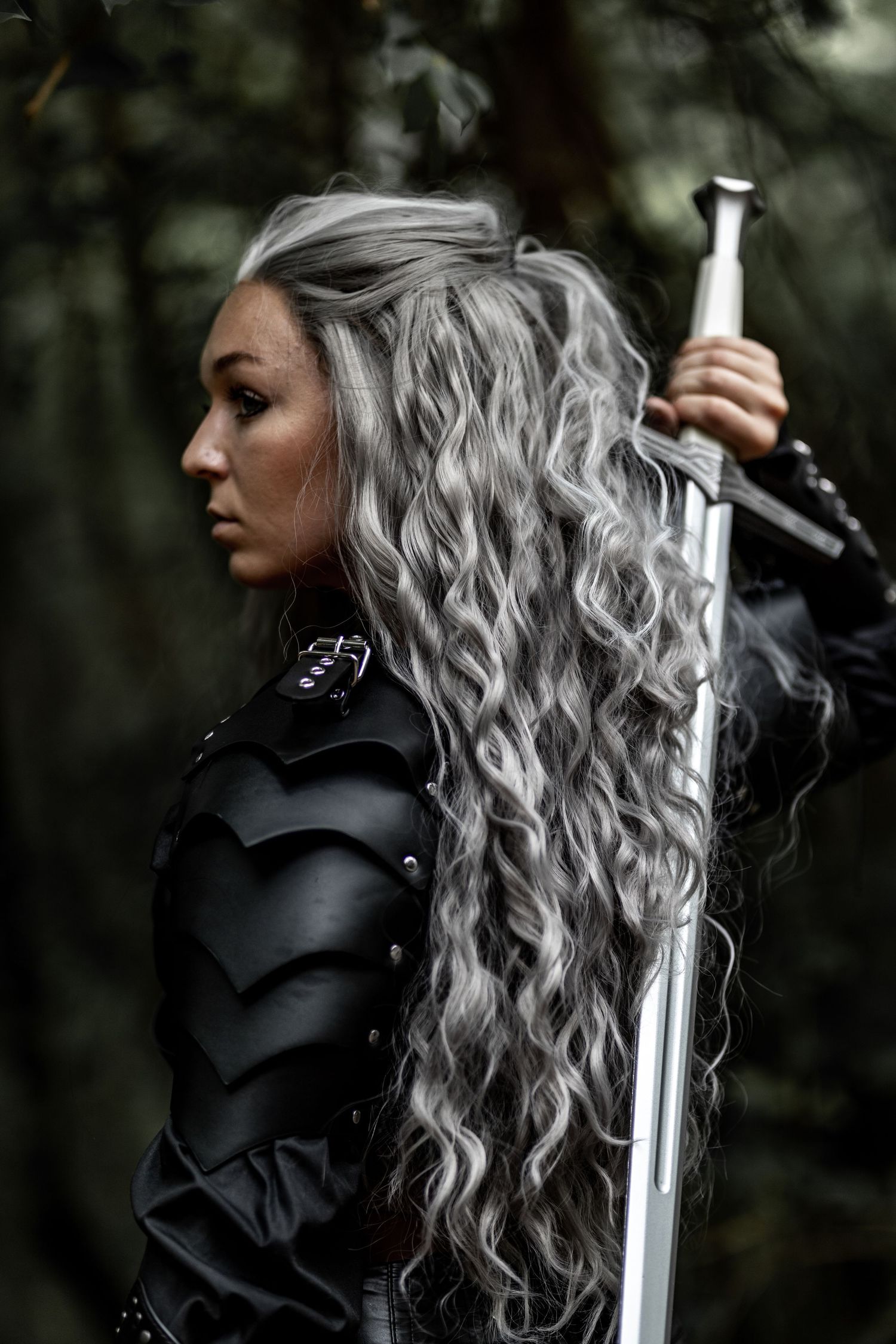 Woman with silver hair in black armor holding sword in forest setting.