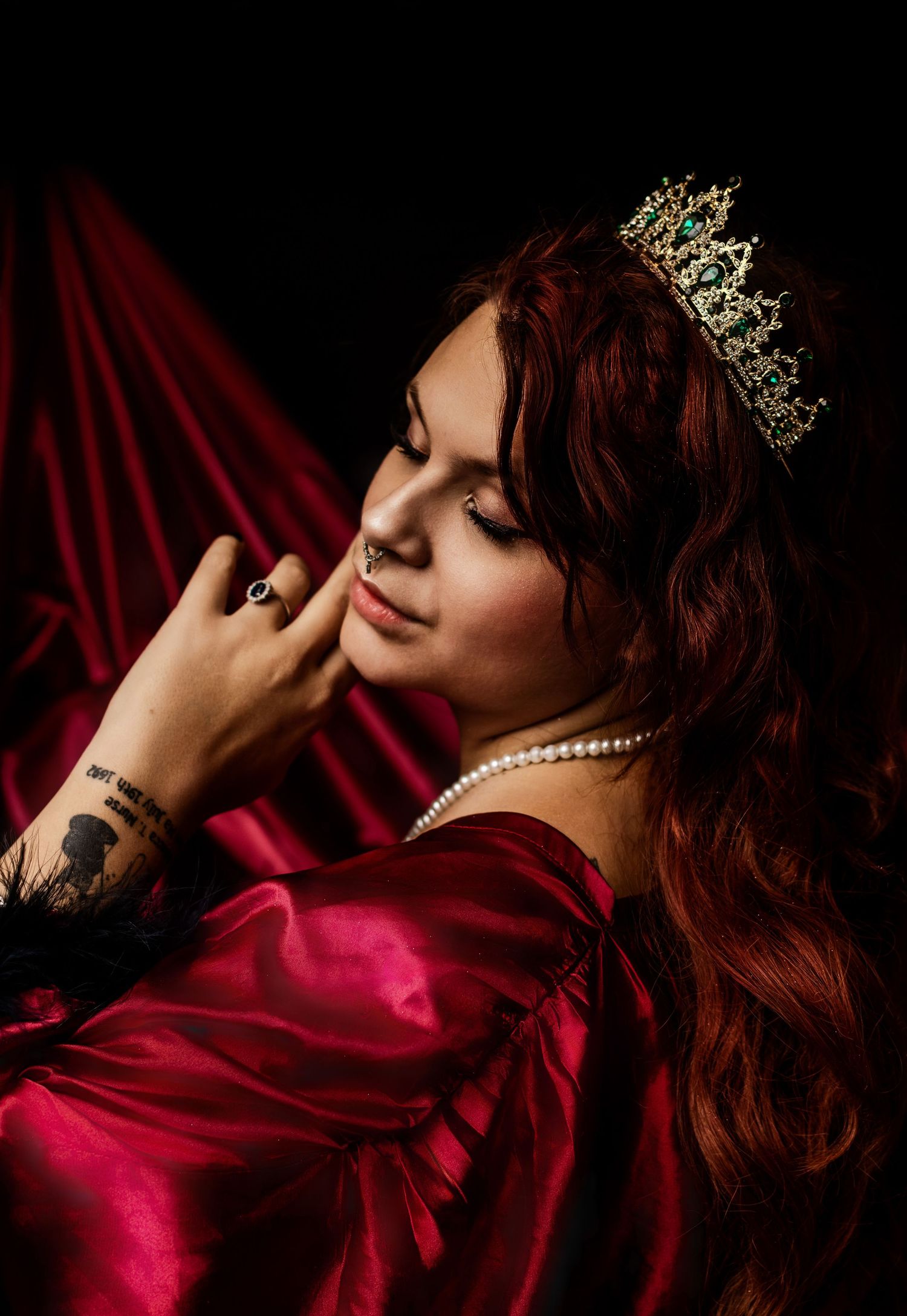 A woman with red hair wearing a gold crown, pearl necklace, and red satin gown poses elegantly against a crimson backdrop.
