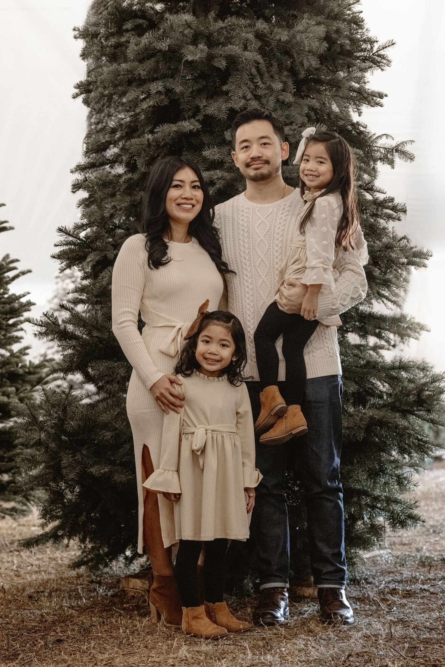 A family poses together in coordinated neutral outfits in front of an evergreen tree.