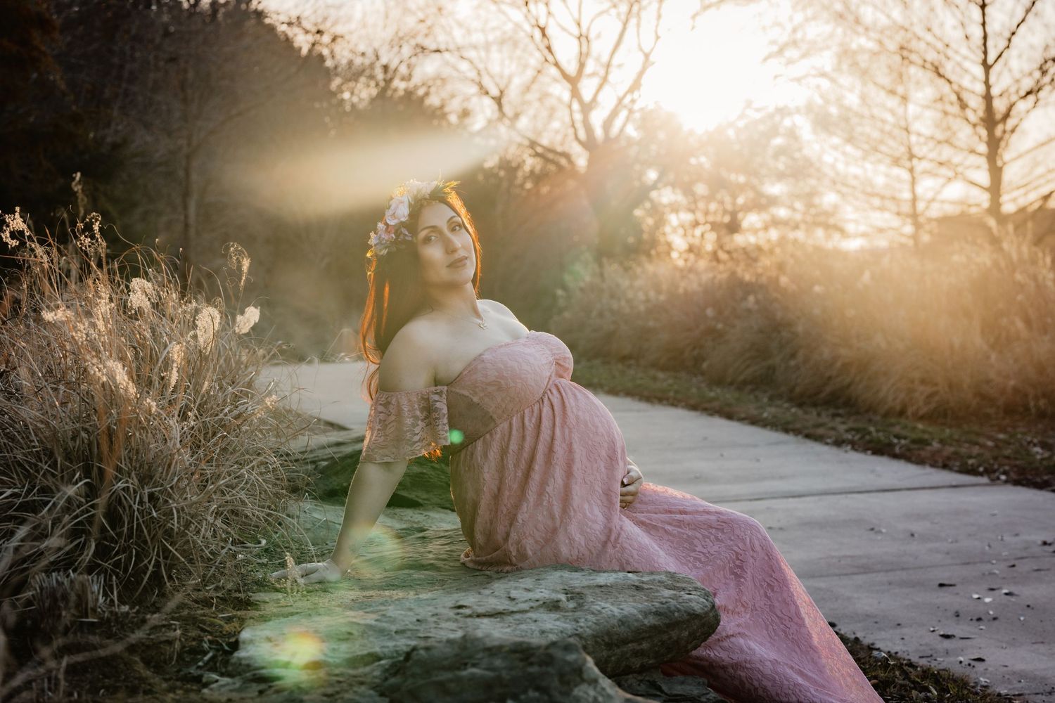 Dreamy sunset portrait of someone in a flowing pink dress reclining on a path surrounded by golden light and tall grass.