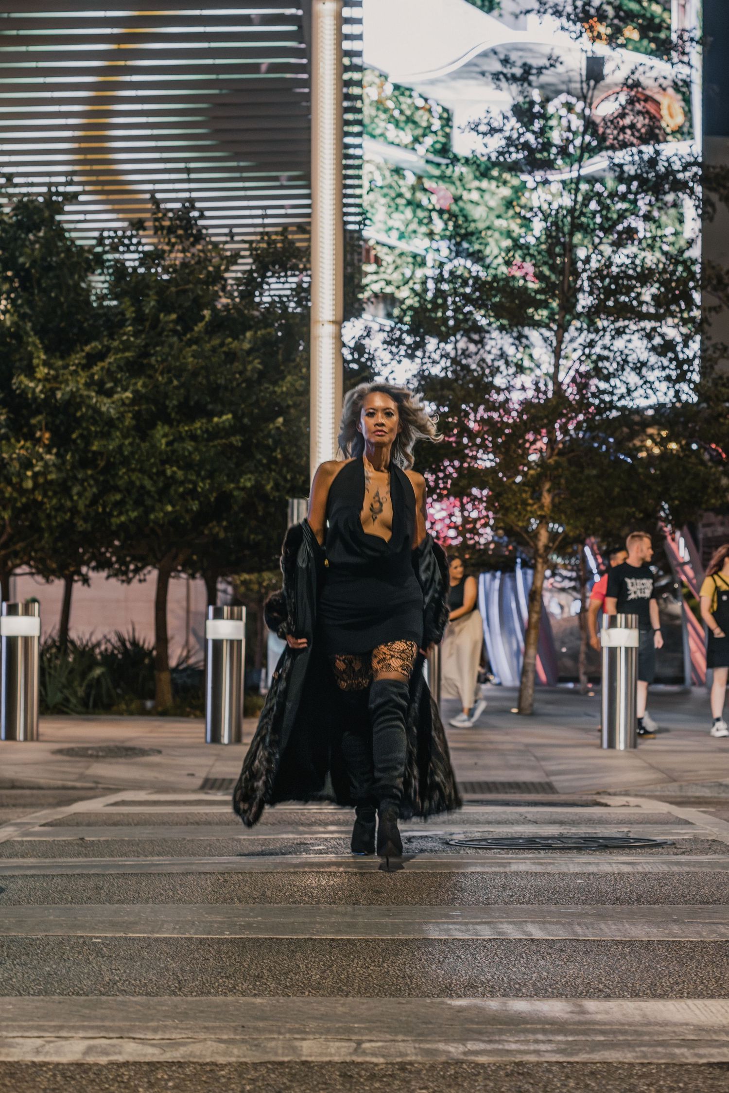 A stylish woman in black outfit with long coat and thigh-high boots walks confidently across an urban crosswalk at night.