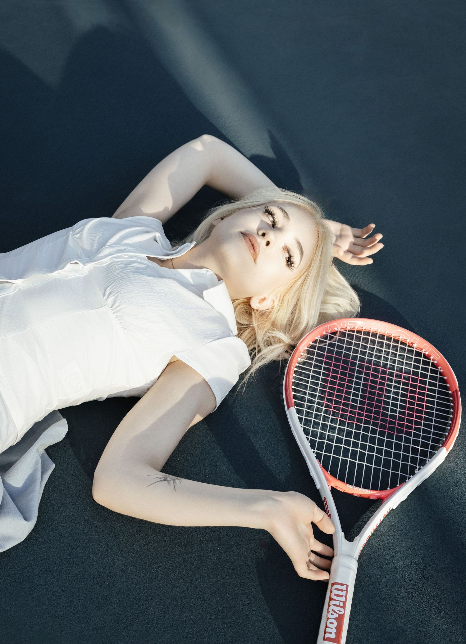 Blonde woman in white dress lying on tennis court holding a Wilson racket in a stylish editorial photo.