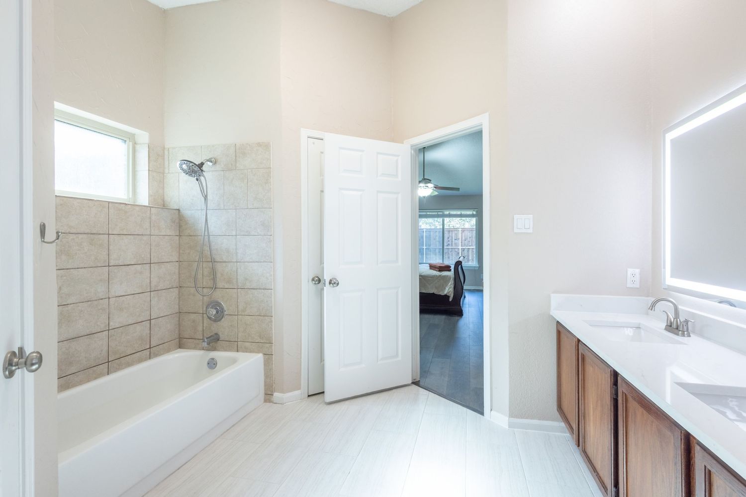 Bright bathroom with tiled tub surround, dual sink vanity, and open door leading to bedroom.