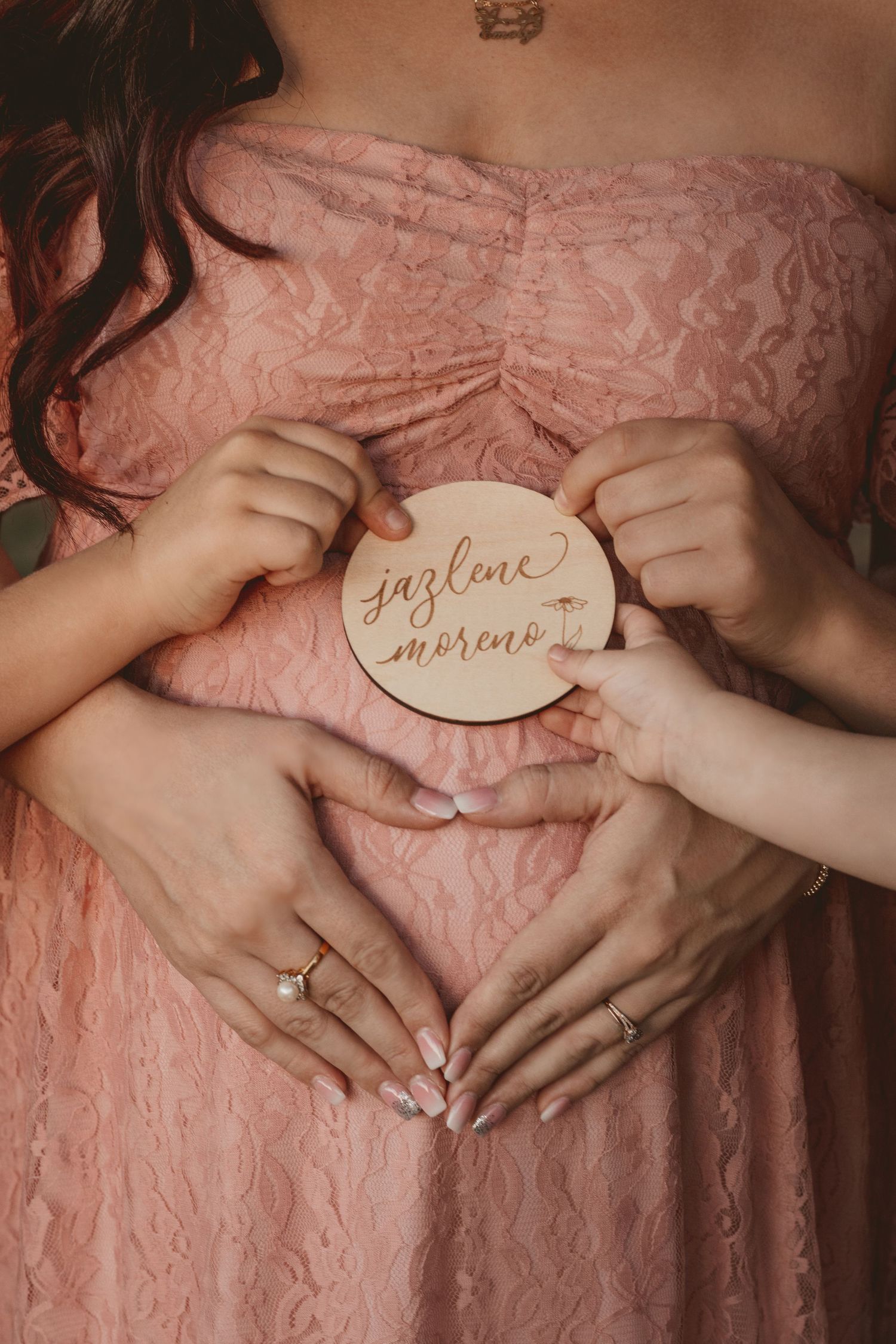 Hands holding wooden signs against a pink lace dress in a maternity photoshoot.