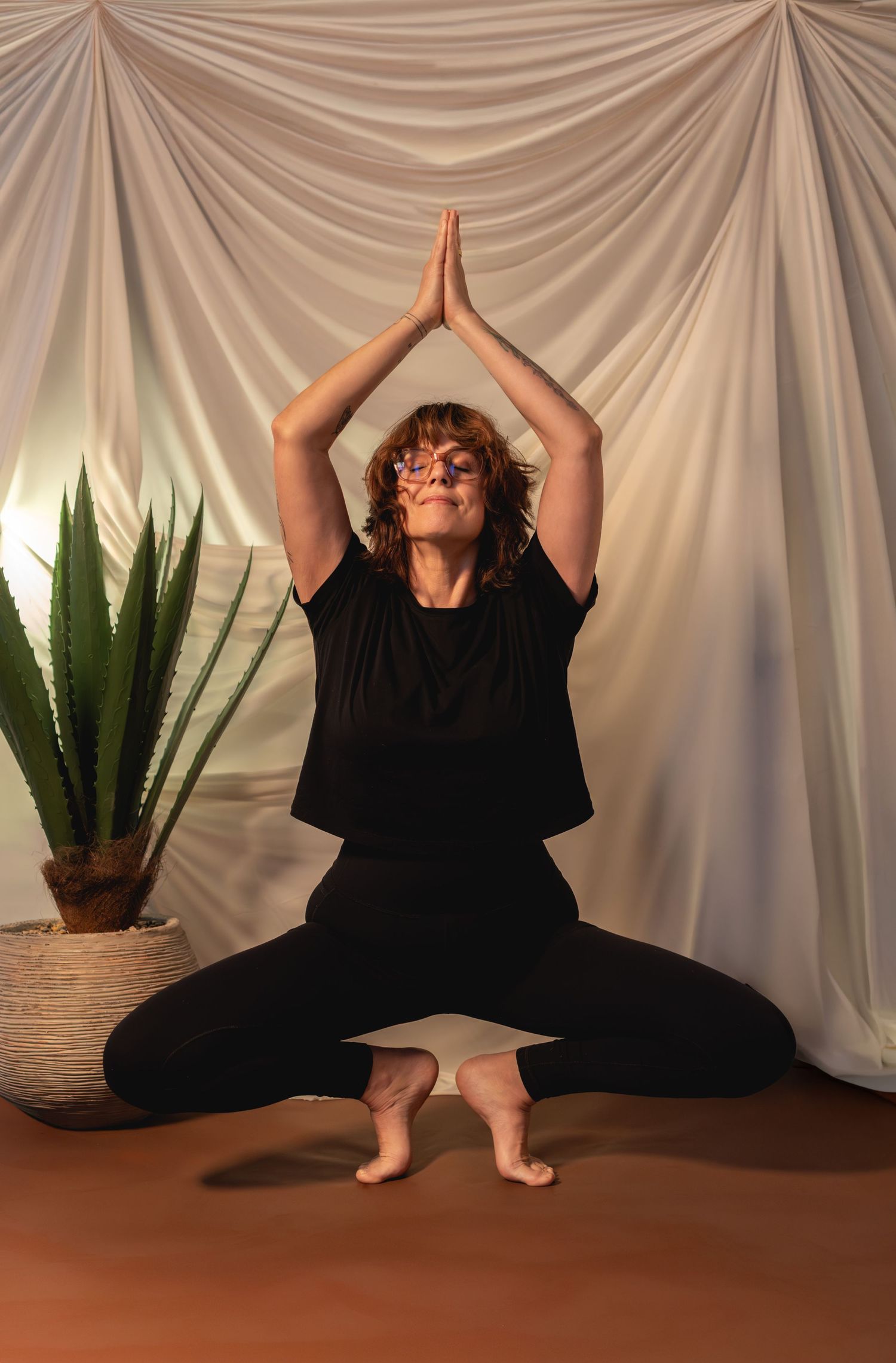 Woman in black outfit performs a seated yoga pose with hands in prayer position overhead against white draped backdrop.