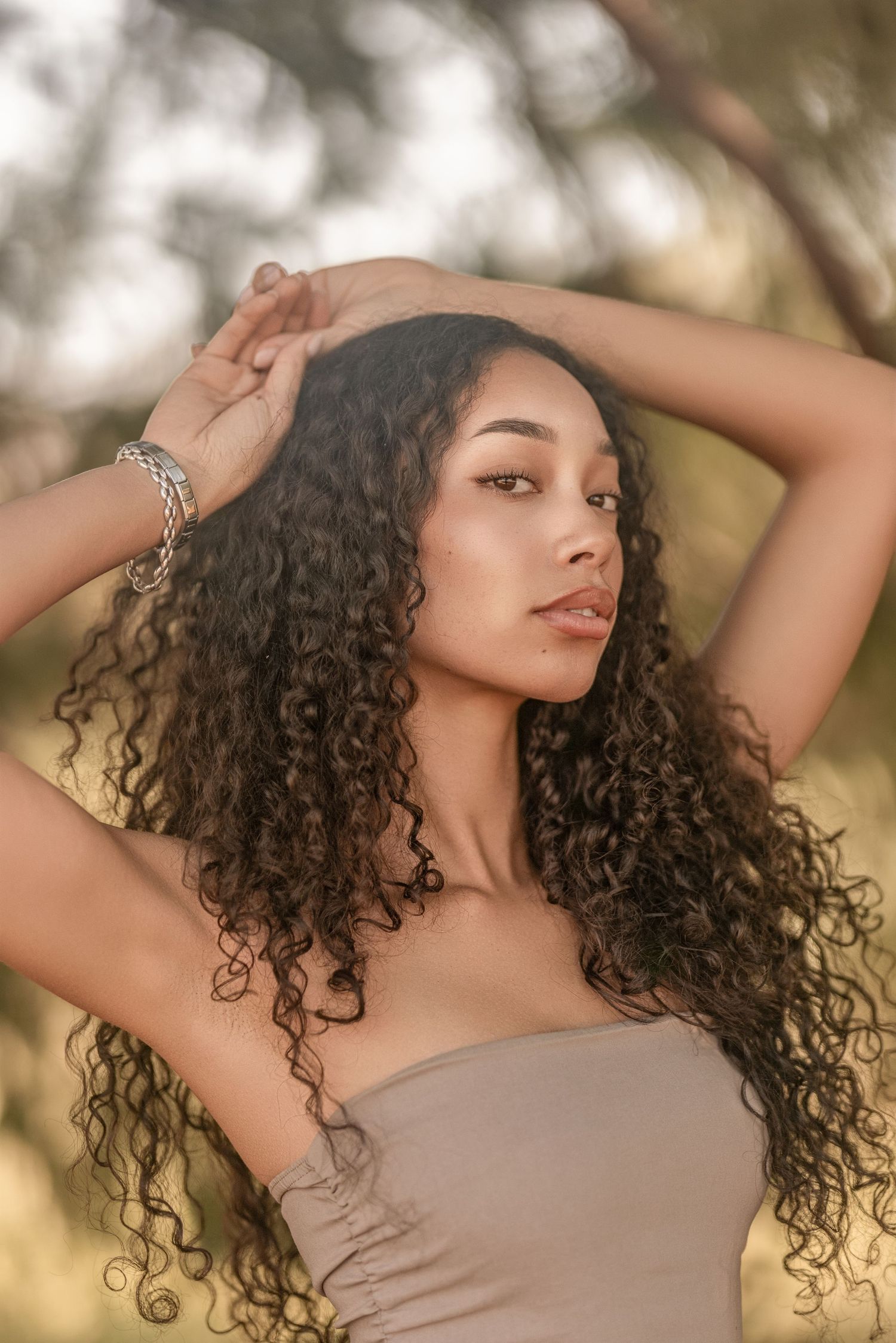 Young woman with long curly hair poses outdoors in a beige top, wearing a silver bracelet, with trees in the background.