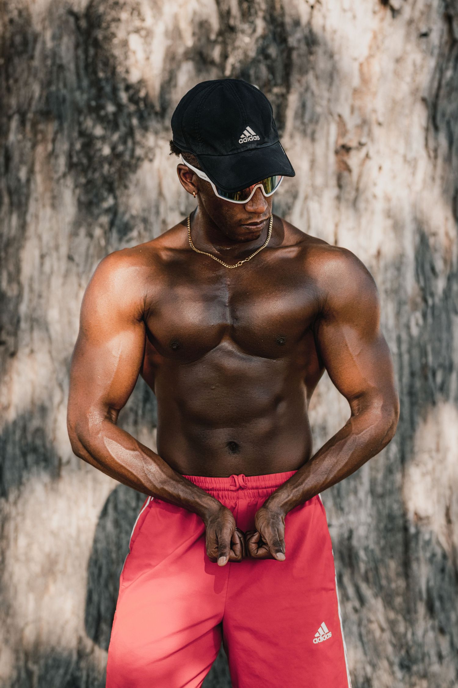 Muscular shirtless Black man wearing Adidas cap, sunglasses, and pink Adidas shorts posing against a textured wall.