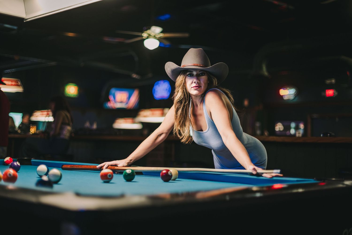 Woman in cowboy hat leaning over a pool table in a dimly lit bar, preparing to take a shot.