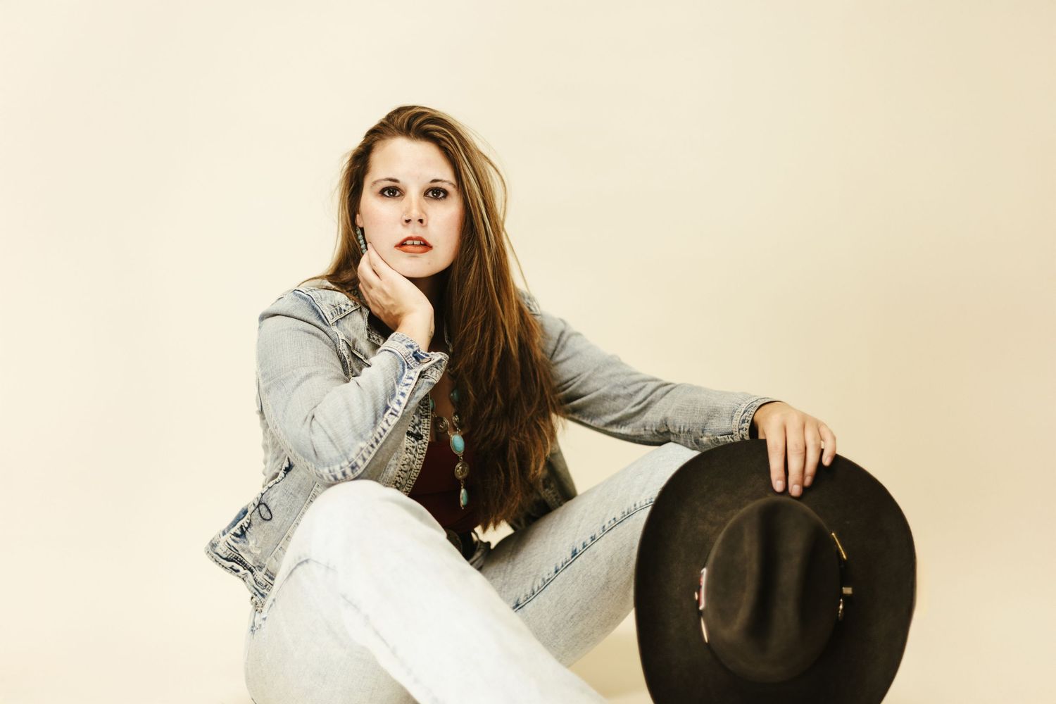 Woman in casual grey outfit sits thoughtfully while holding a black hat against beige background.