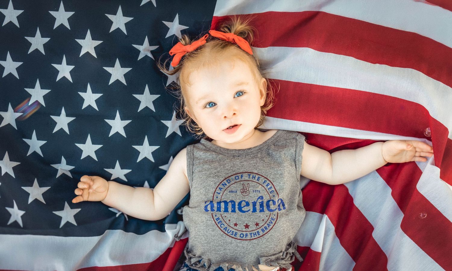 Baby girl in gray America shirt lying on US flag with red bow, perfect for patriotic Fourth of July celebration.