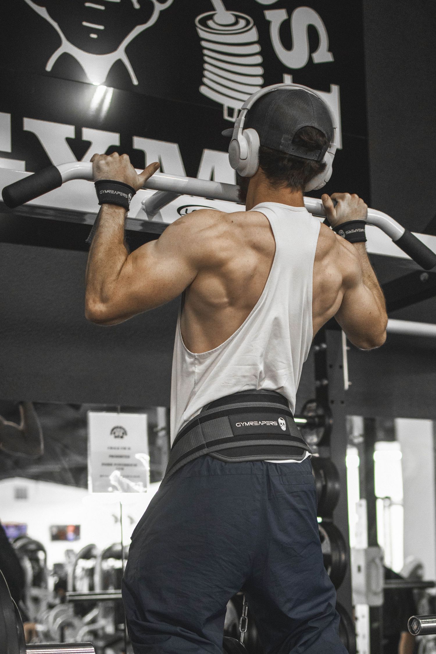 Muscular man doing pull-ups at the gym wearing headphones, a white tank top, and a weightlifting belt.