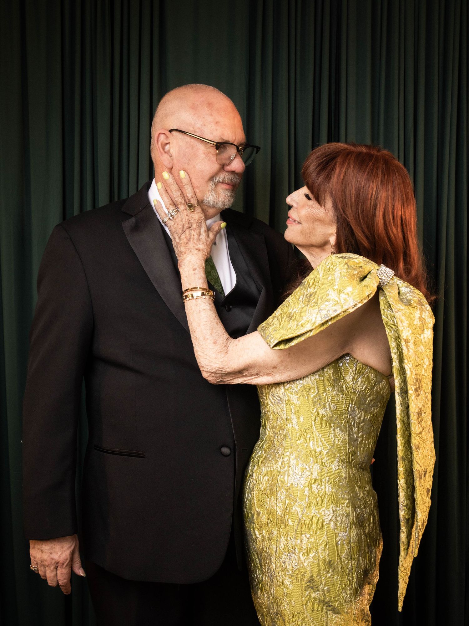 Elegant couple in formal attire, man in black tuxedo and woman in gold gown, sharing a romantic moment.